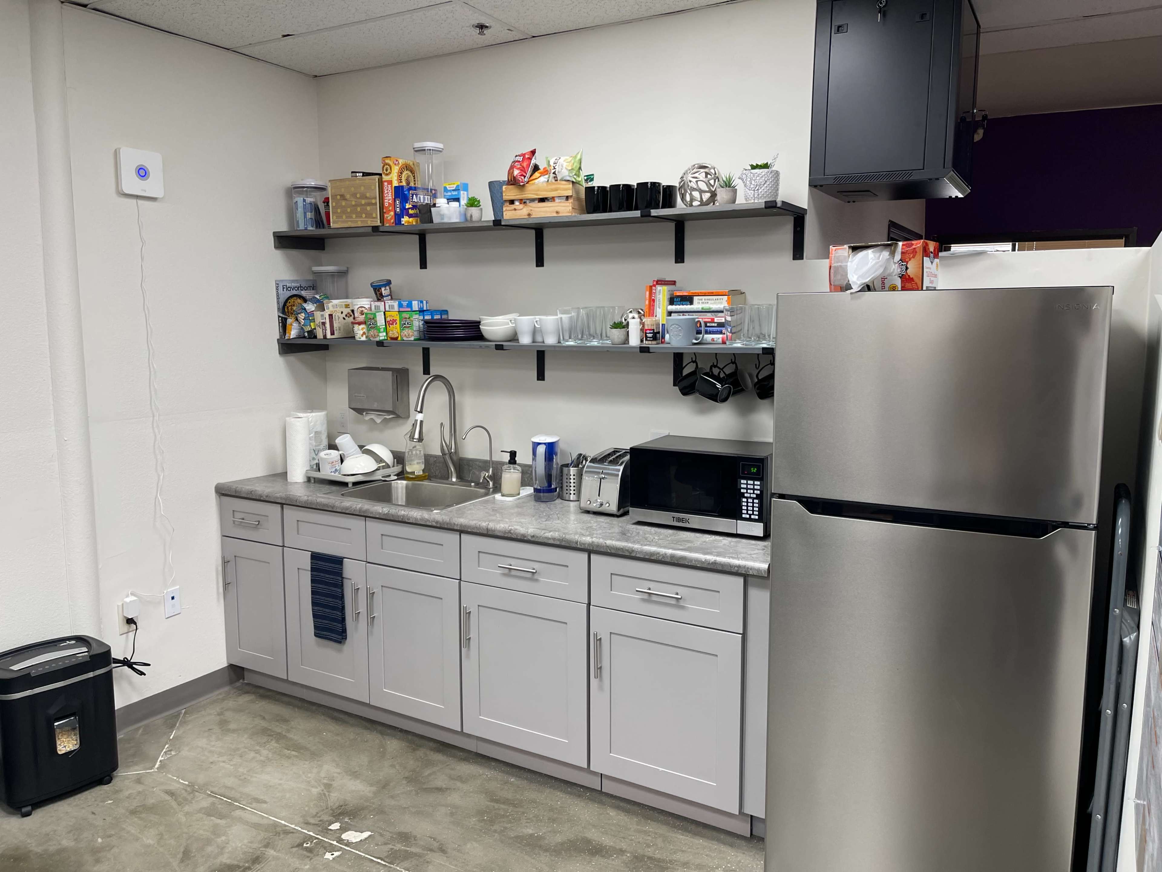 A kitchen area with gray cabinets, a stainless steel refrigerator, a microwave, and open shelves stocked with various food items and dishware.