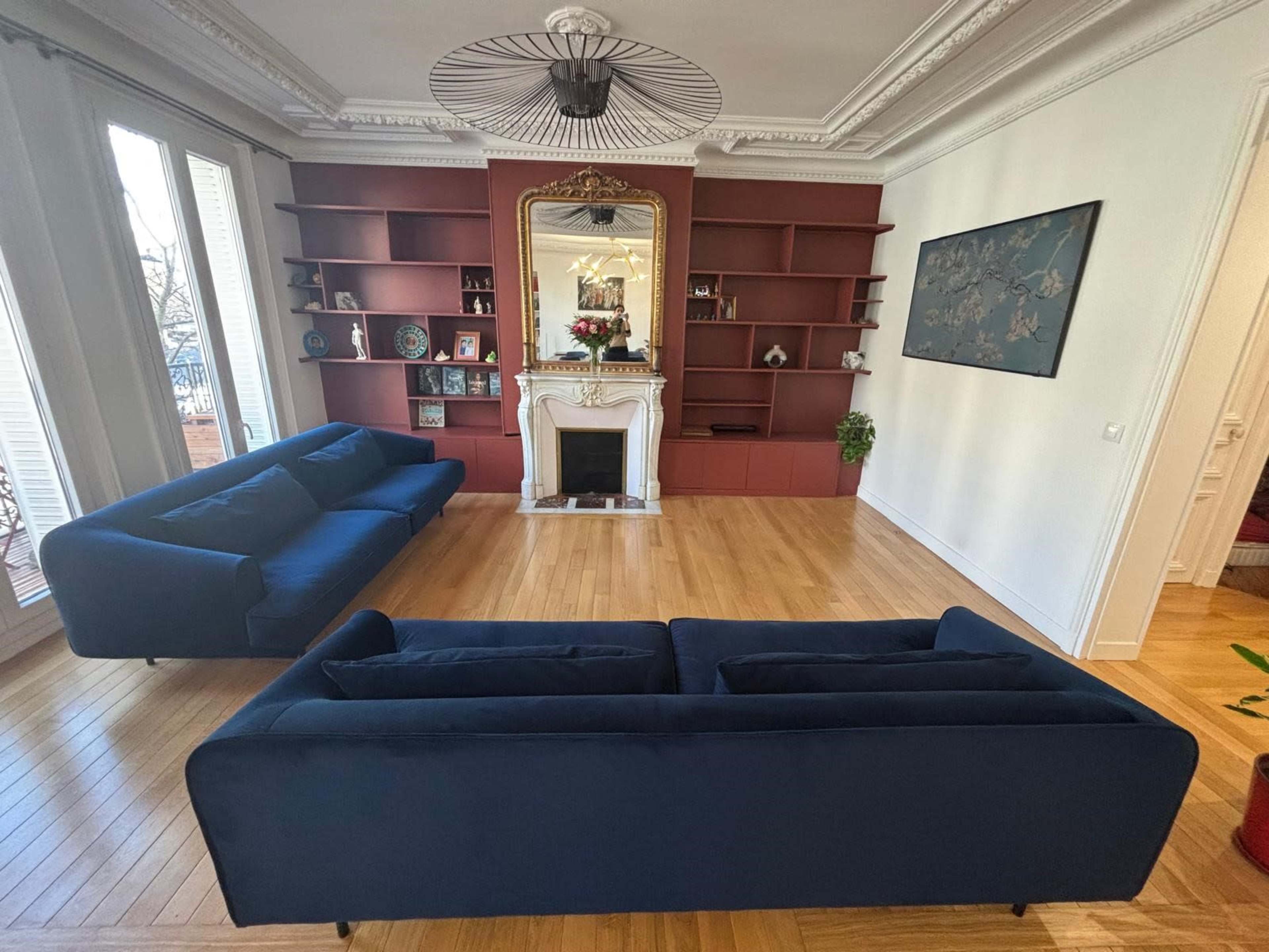 The room features two blue sofas arranged around a white fireplace, with a red bookshelf and decorative items lining the walls.