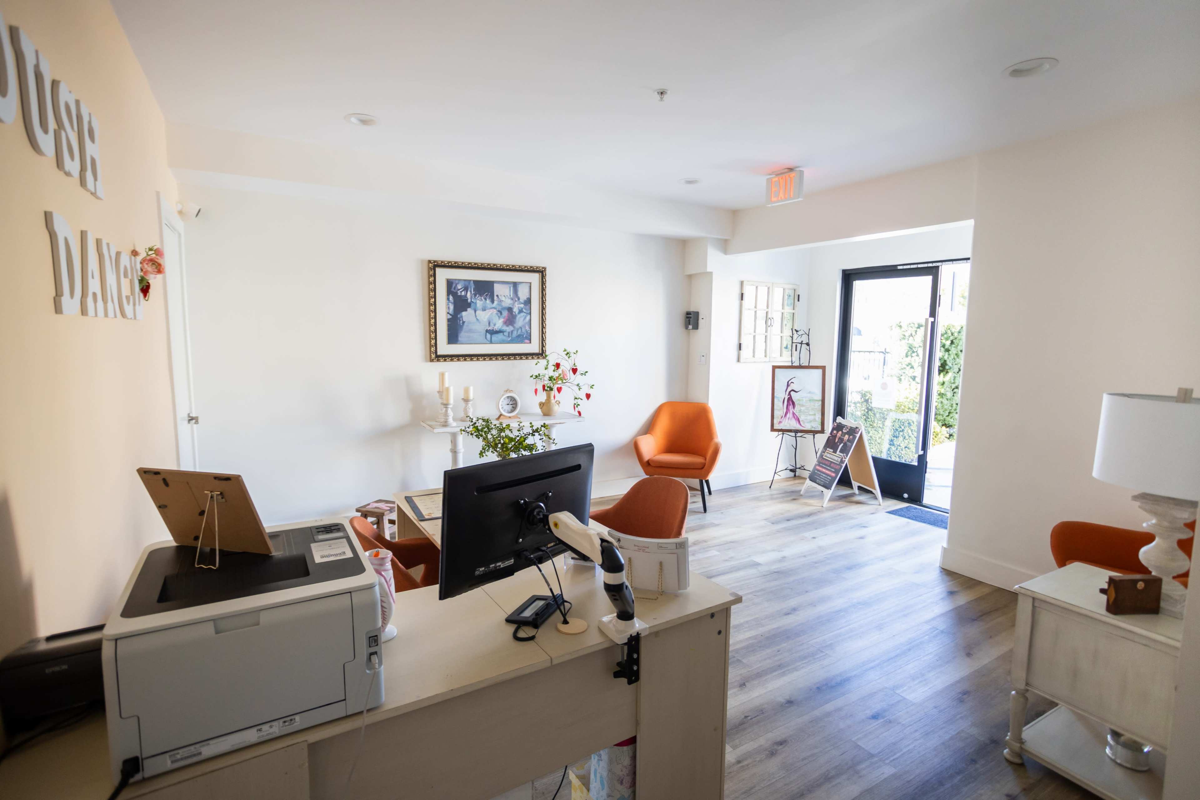 A reception area with a printer, a desk computer, and orange chairs near a doorway.