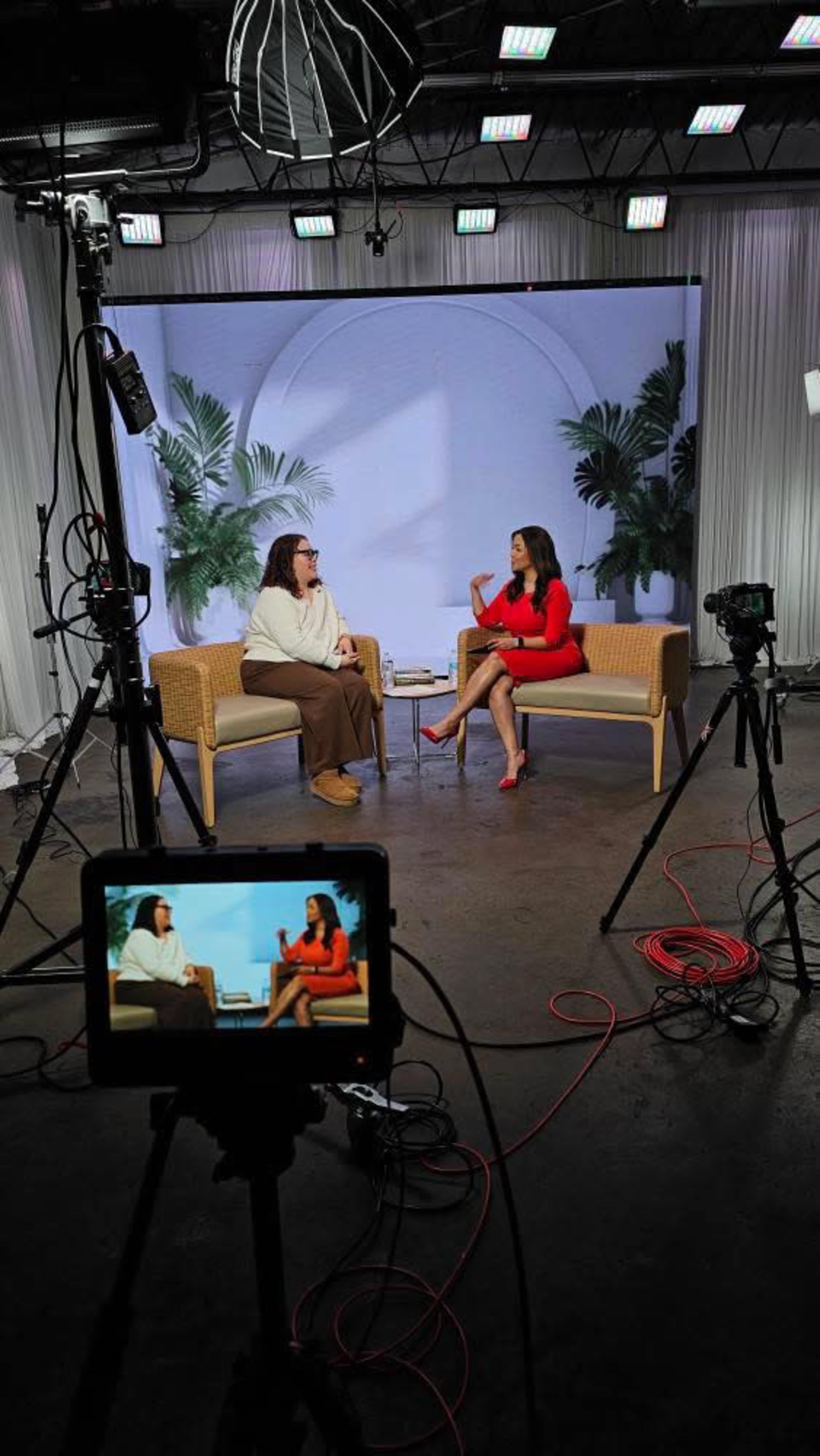 Two women are seated on a set designed for an interview, with cameras and lights positioned around them.