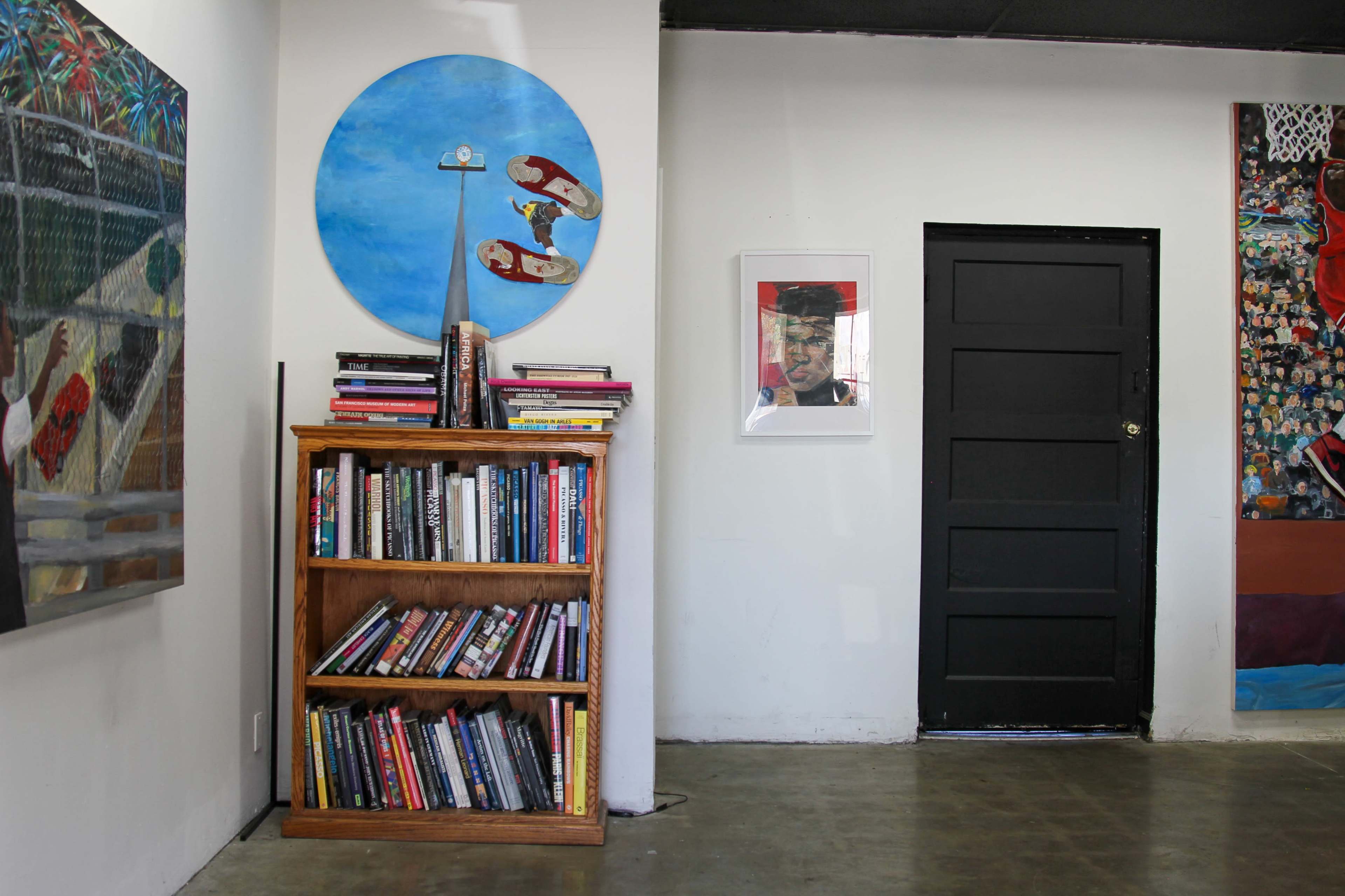 The scene features a wooden bookshelf filled with books against a wall decorated with colorful artwork, including a round blue painting and a framed portrait.