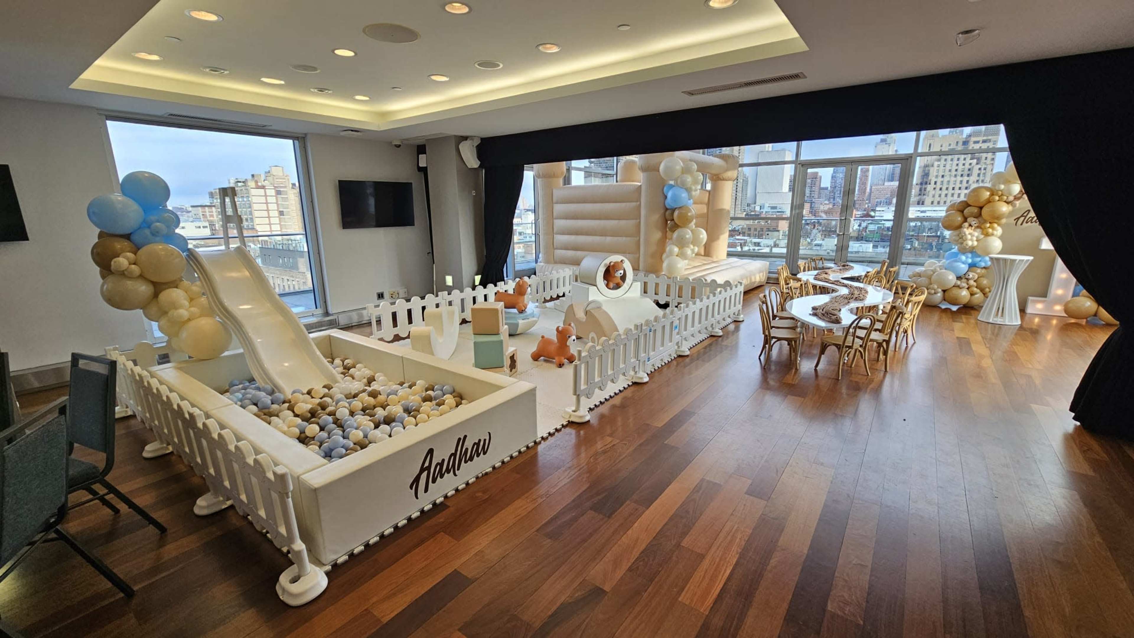 The image shows a decorated event space featuring a play area with a slide, a ball pit, and a table set for a celebration surrounded by balloon arrangements.