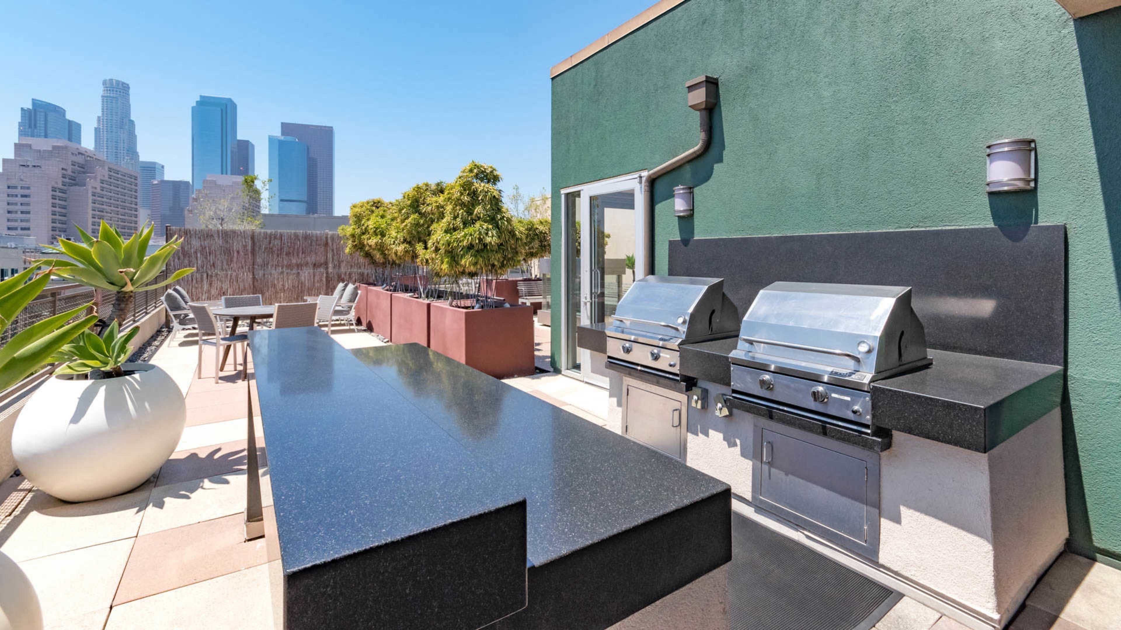An outdoor kitchen area with two stainless steel grills and a long black countertop, set against a backdrop of downtown Los Angeles skyscrapers.