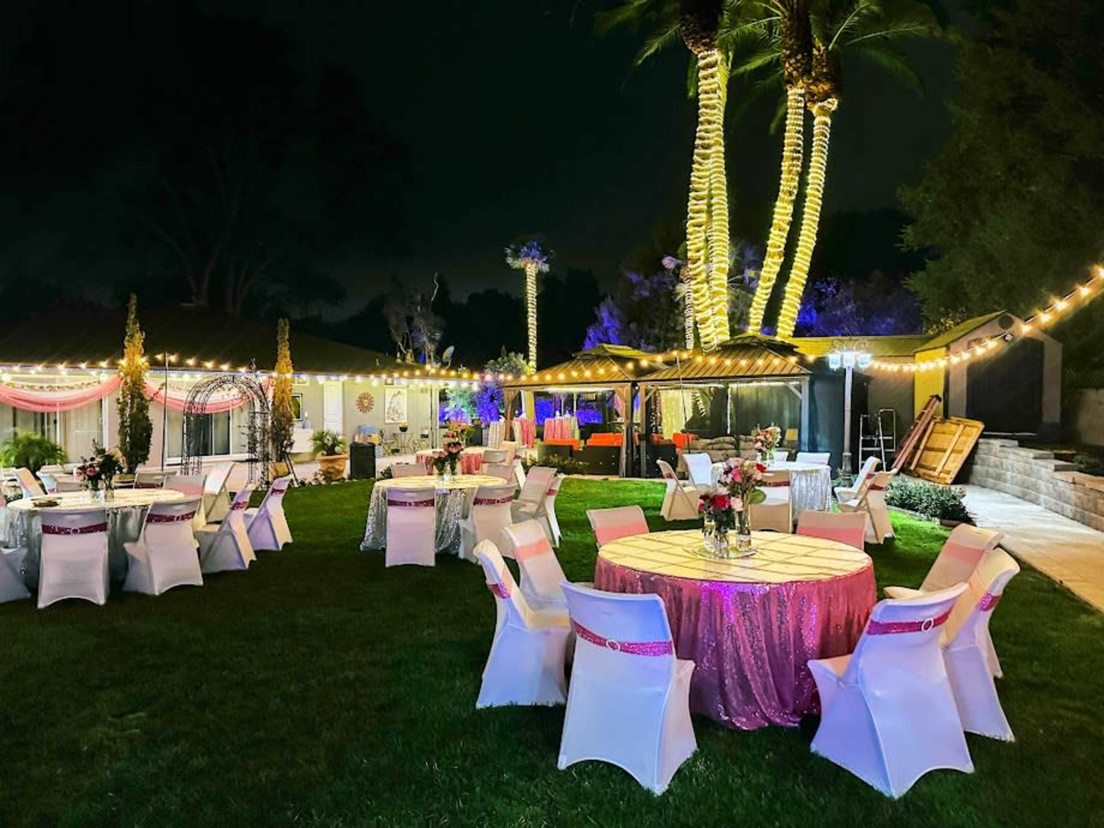 A decorated outdoor venue features round tables covered in bright tablecloths and surrounded by white chairs, illuminated by string lights and palm trees.