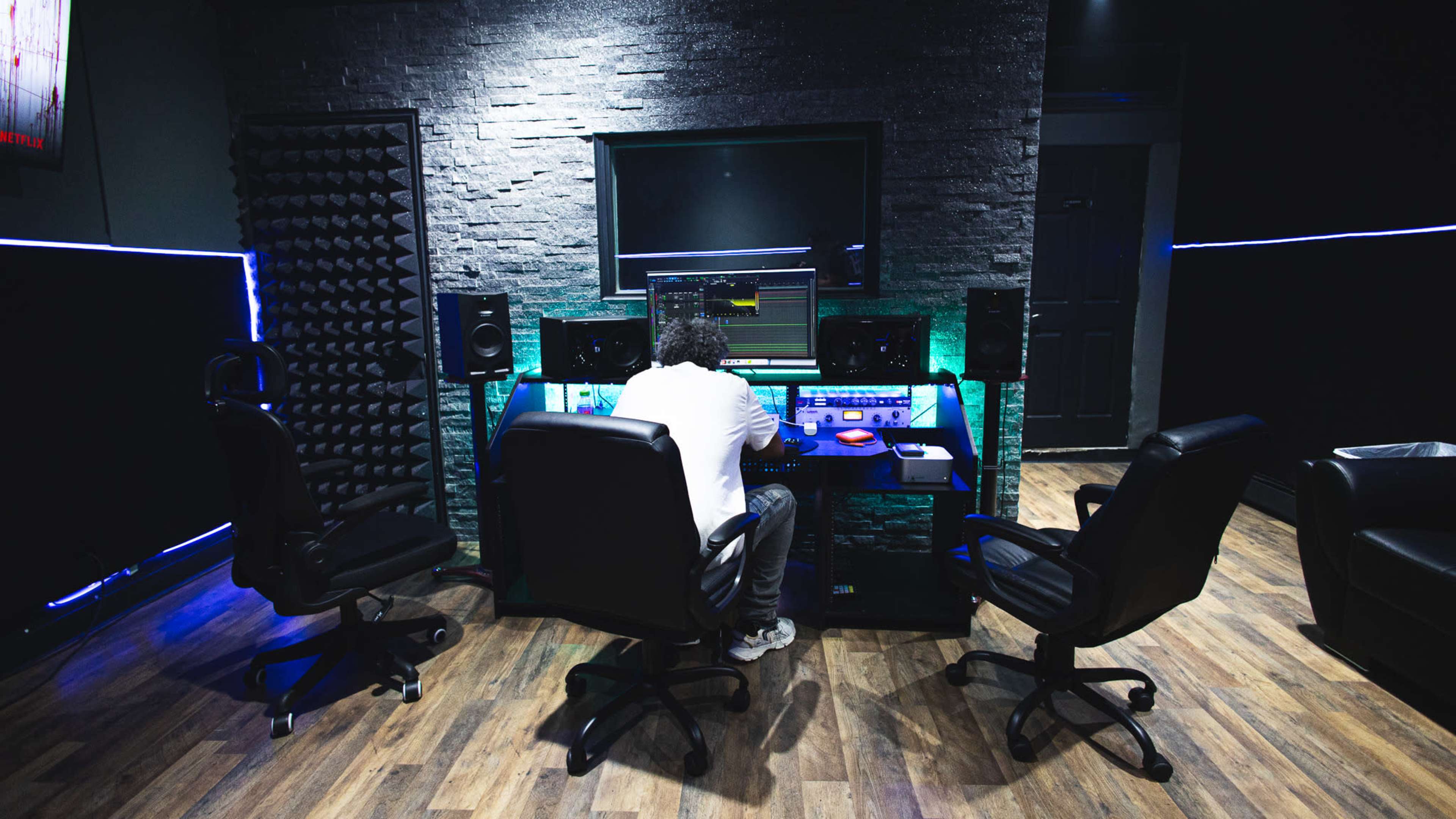 A person is seated at a mixing console in a recording studio, surrounded by speakers and acoustic panels.