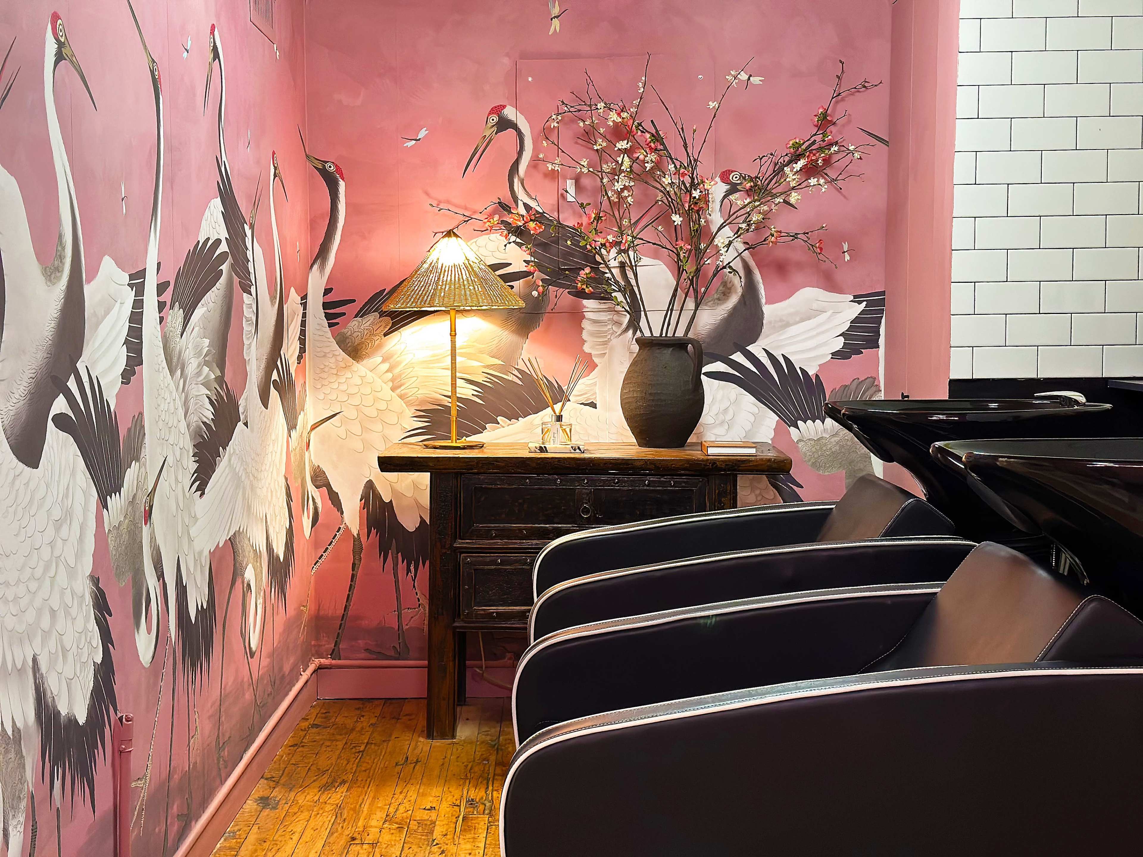 A cozy salon interior with a pink wall mural of cranes, a wooden table with a lamp, a vase with branches, and black salon chairs.