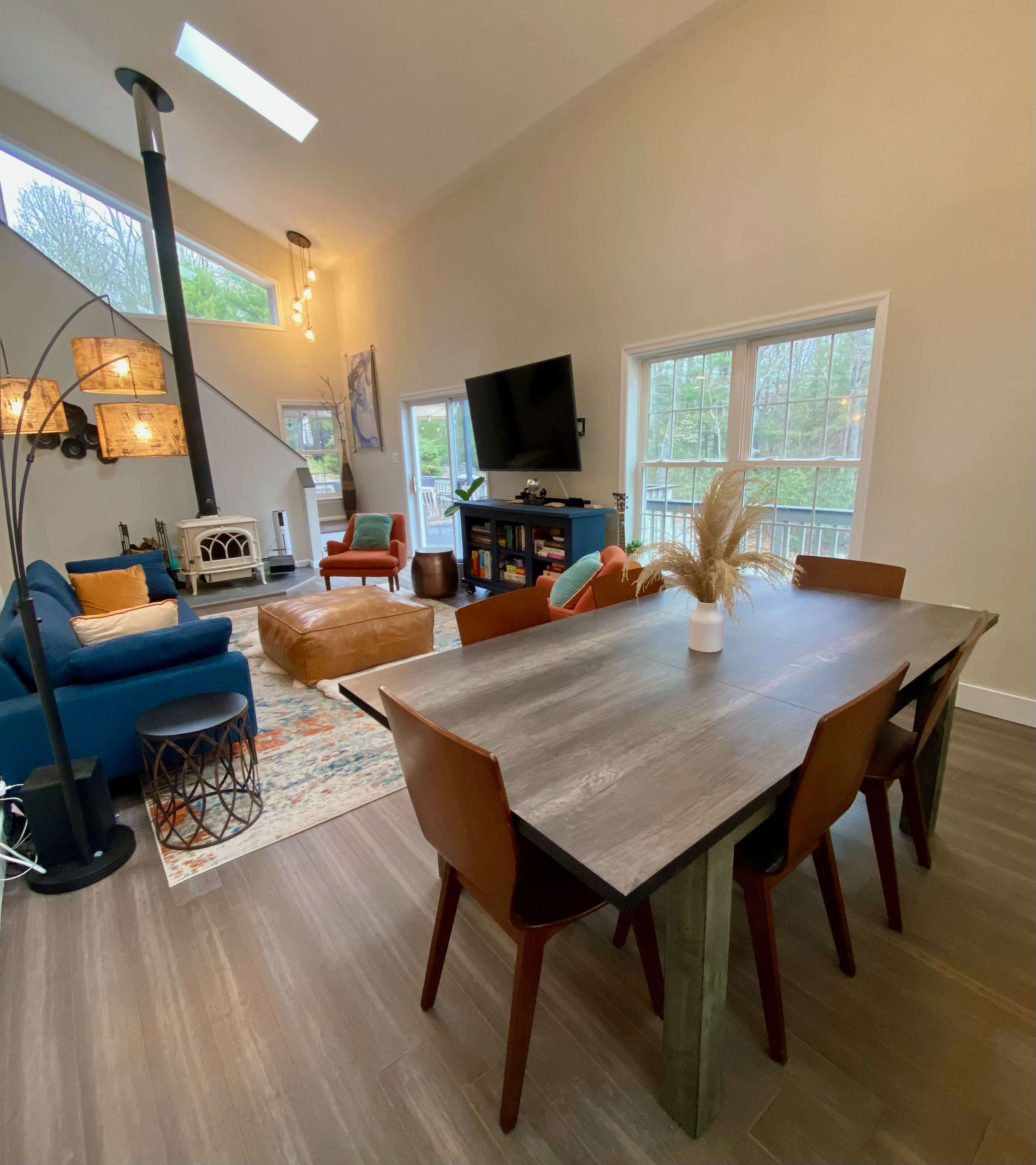 A dining table with brown chairs is positioned in a bright, open living area that features a blue couch, a television, and large windows.