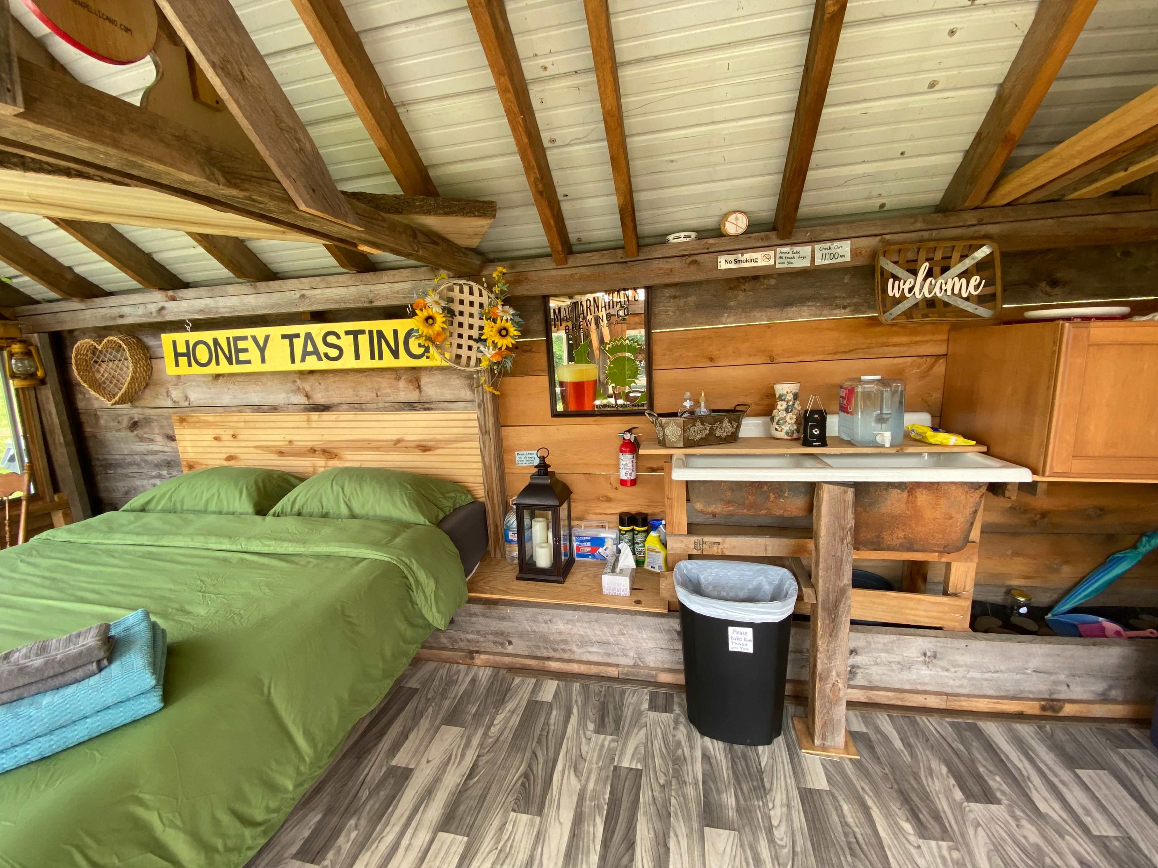 The image shows a rustic interior of a small cabin featuring a bed, a kitchenette area, and decorative signage.