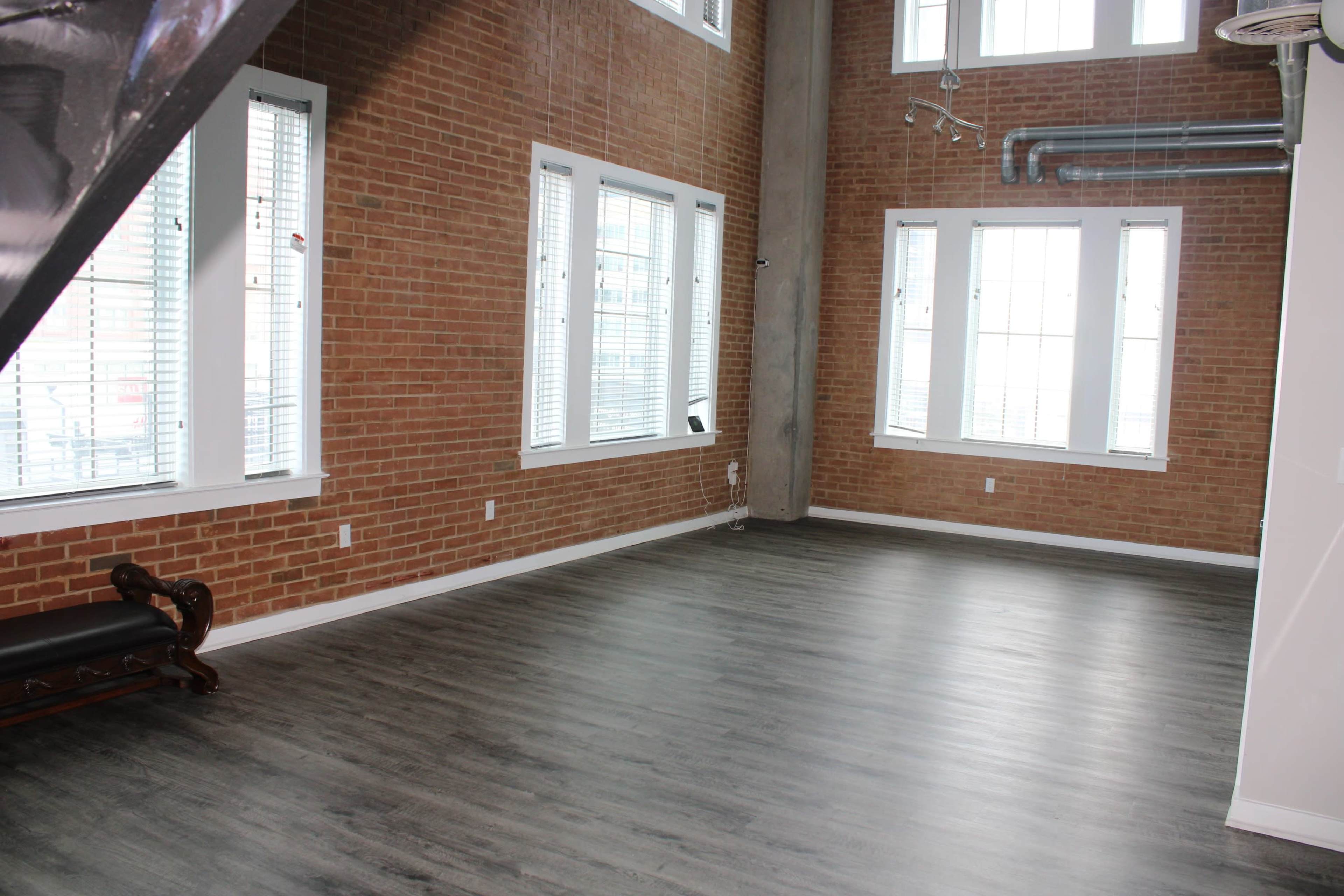 An interior space with exposed brick walls, large windows, and a wooden floor.
