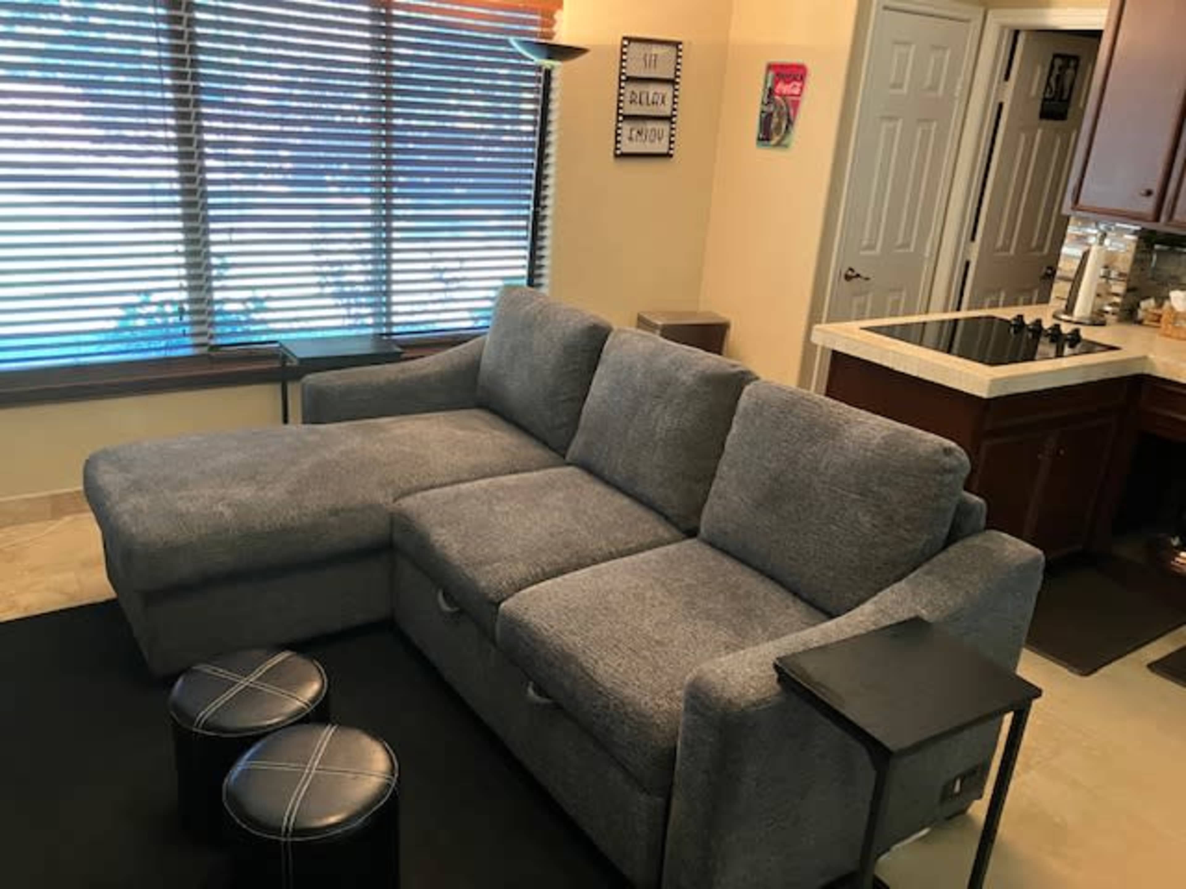 A gray sectional sofa is positioned in a room with large windows covered by wooden blinds, accompanied by two small black ottomans and a kitchen area in the background.