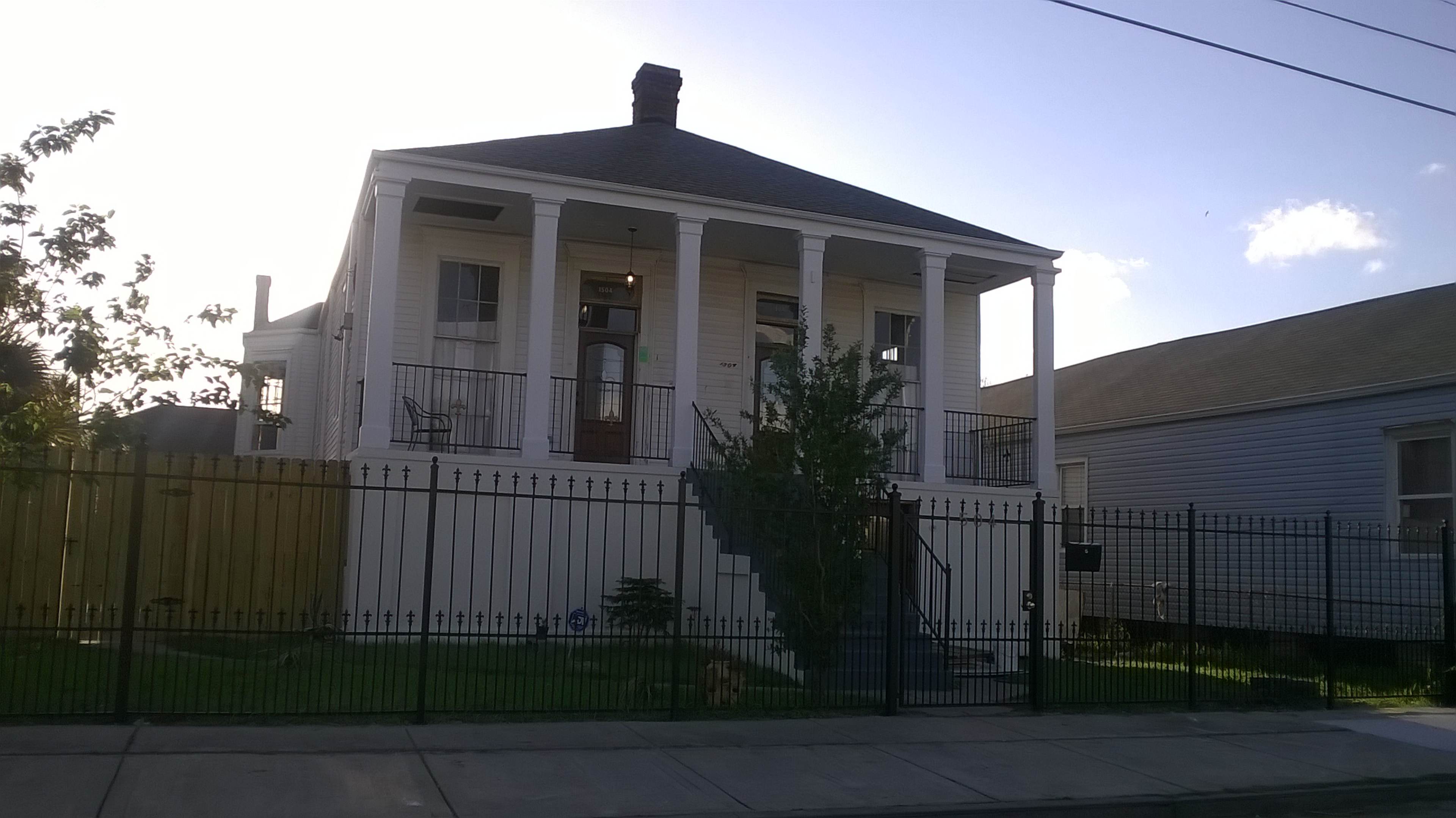 A two-story white house with a front porch supported by four columns, surrounded by a black wrought-iron fence.