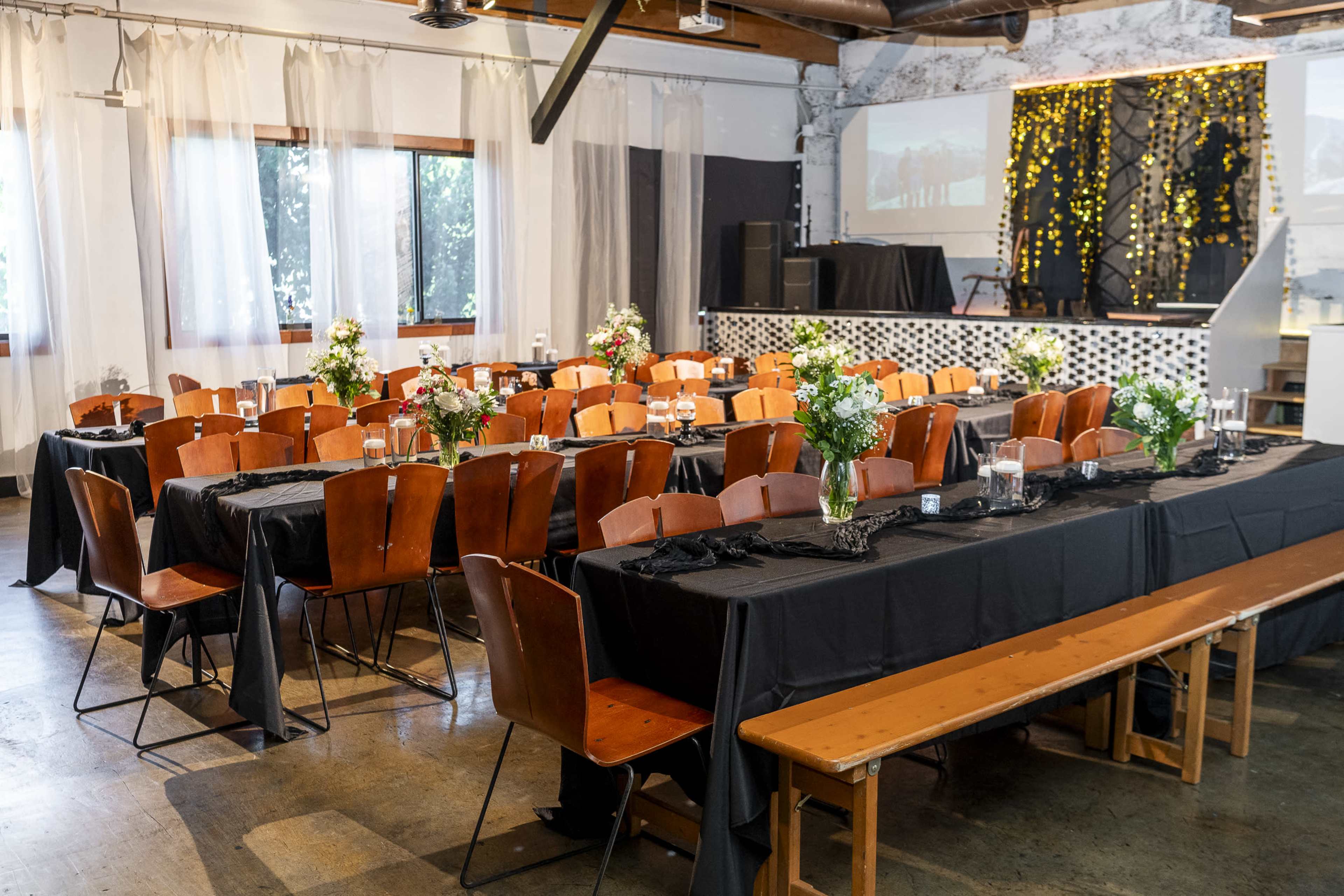 A spacious event venue is set up with rows of tables covered in black tablecloths and adorned with floral centerpieces, surrounded by wooden chairs and benches.