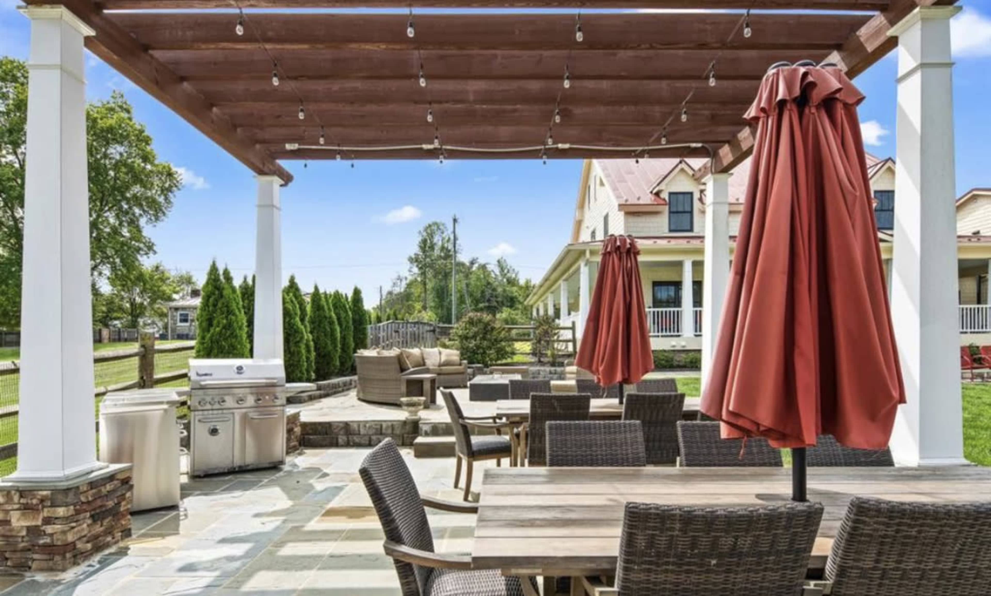 The image shows an outdoor patio with a wooden pergola, seating arrangements, and a grill, surrounded by well-maintained greenery.