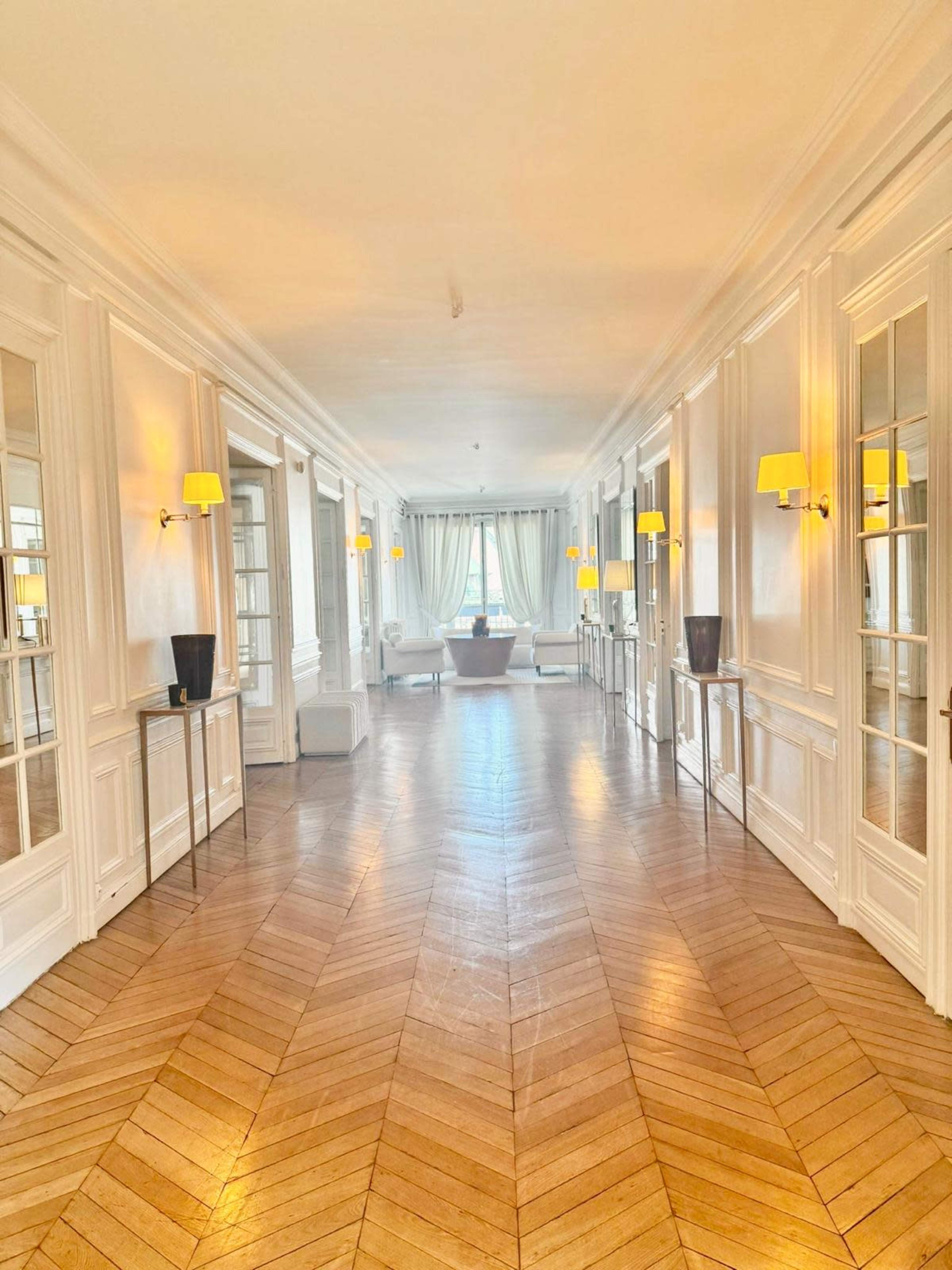 The image shows a long hallway with a herringbone wood floor, white walls, and yellow wall sconces, leading to a bright area with large windows at the end.