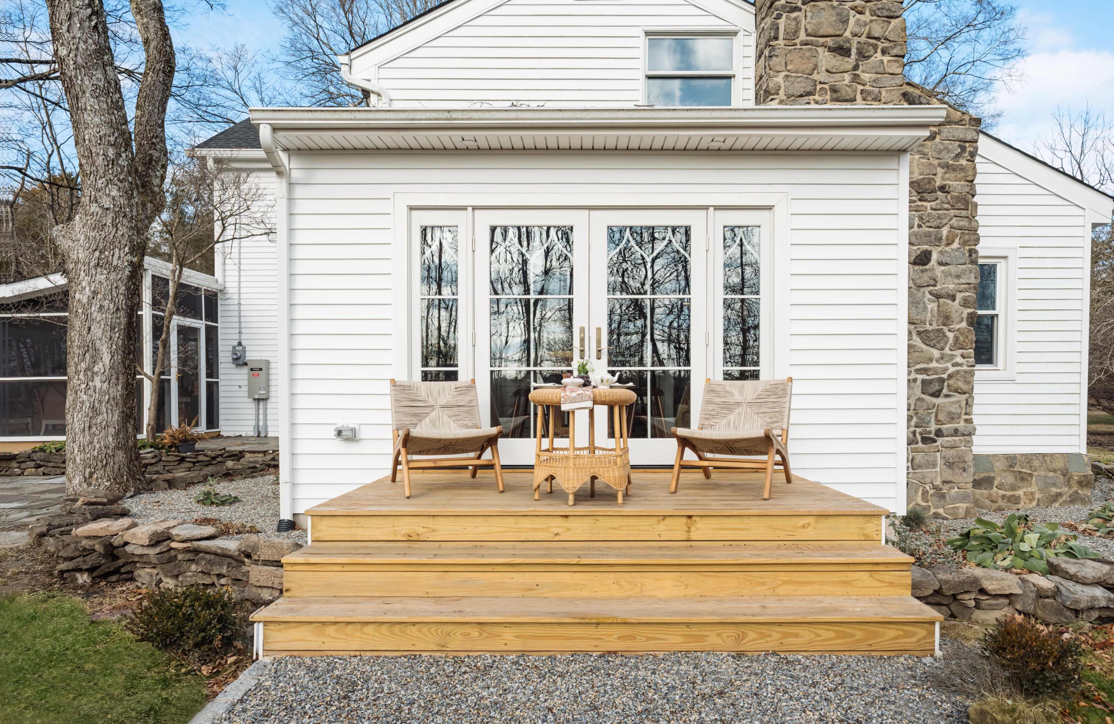 A wooden deck with two chairs and a small table is located at the back of a house, surrounded by trees and a stone wall.