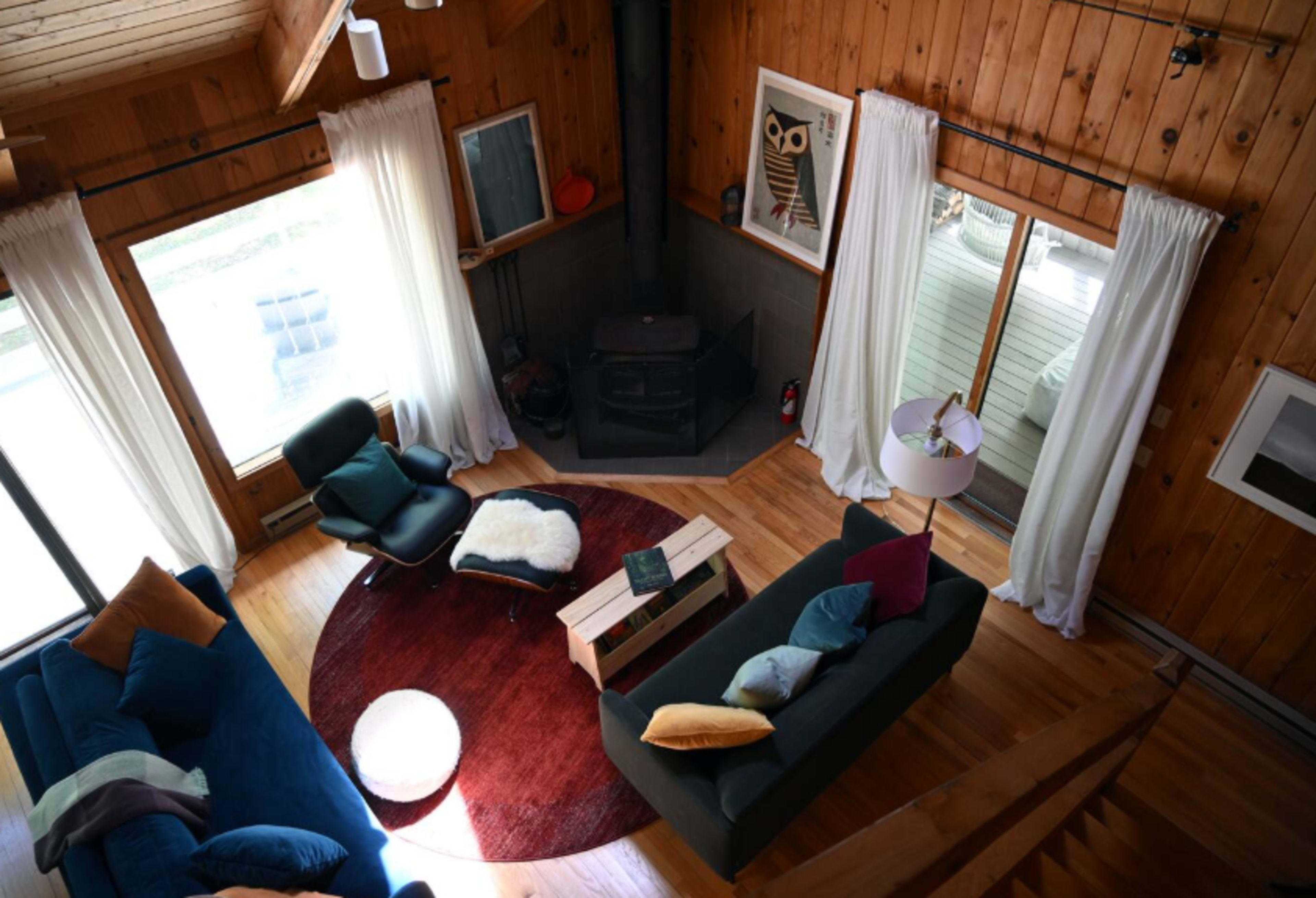 The image shows a cozy living room with a circular rug, a wood-burning stove, large windows, and a mix of modern and rustic furniture.