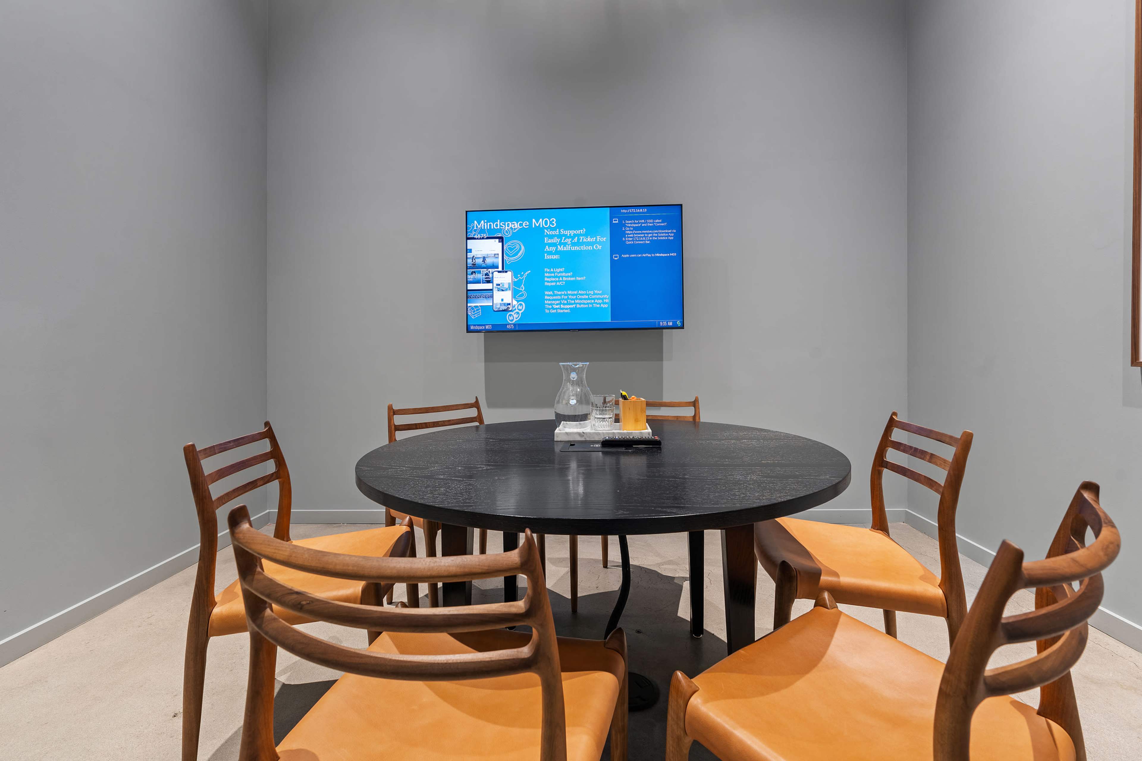 A conference room features a circular black table surrounded by wooden chairs and a wall-mounted screen displaying information.