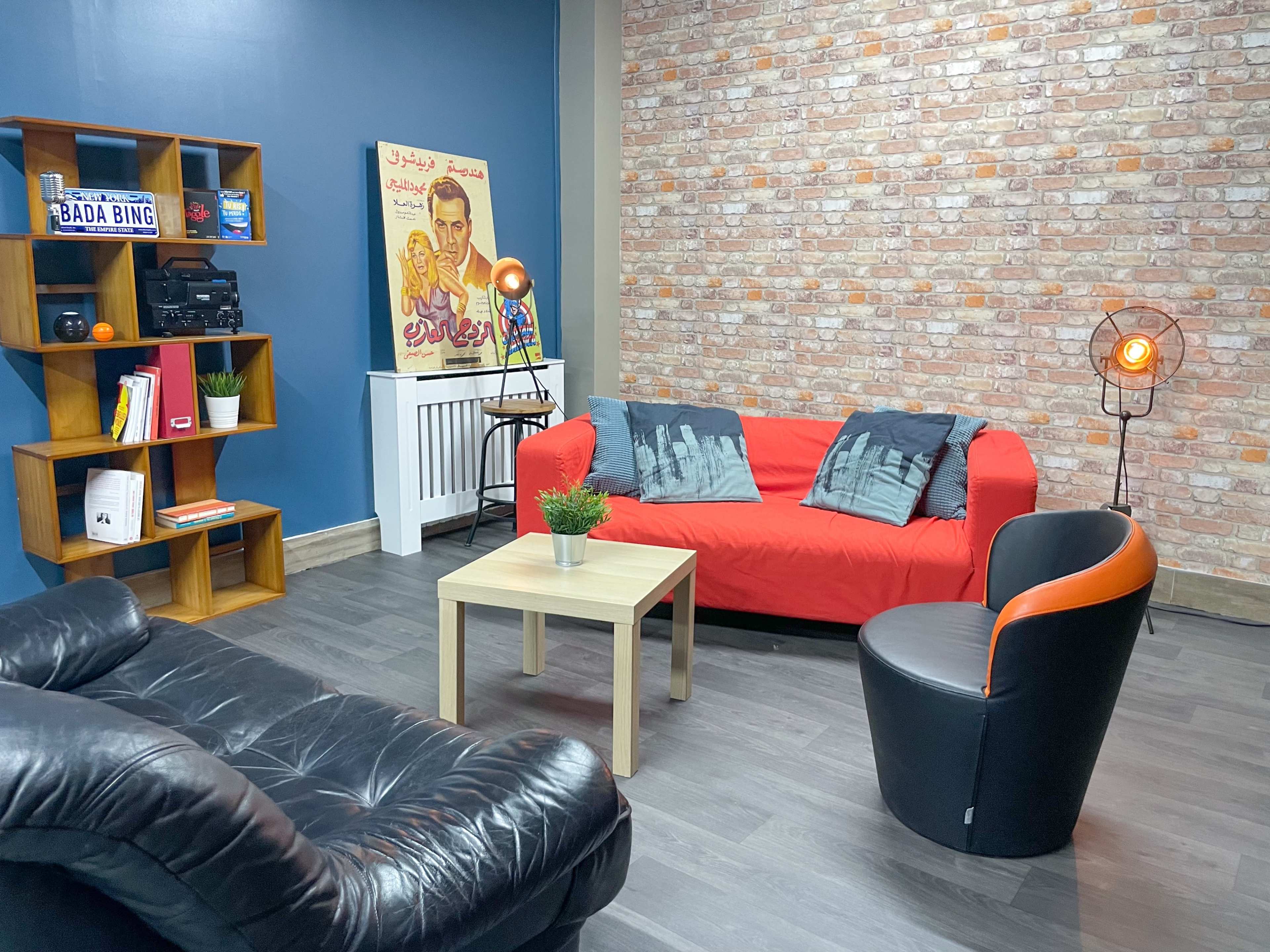A cozy living room with a red couch, a black leather chair, a small beige coffee table, and a decorative bookshelf against a blue wall with a brick accent.