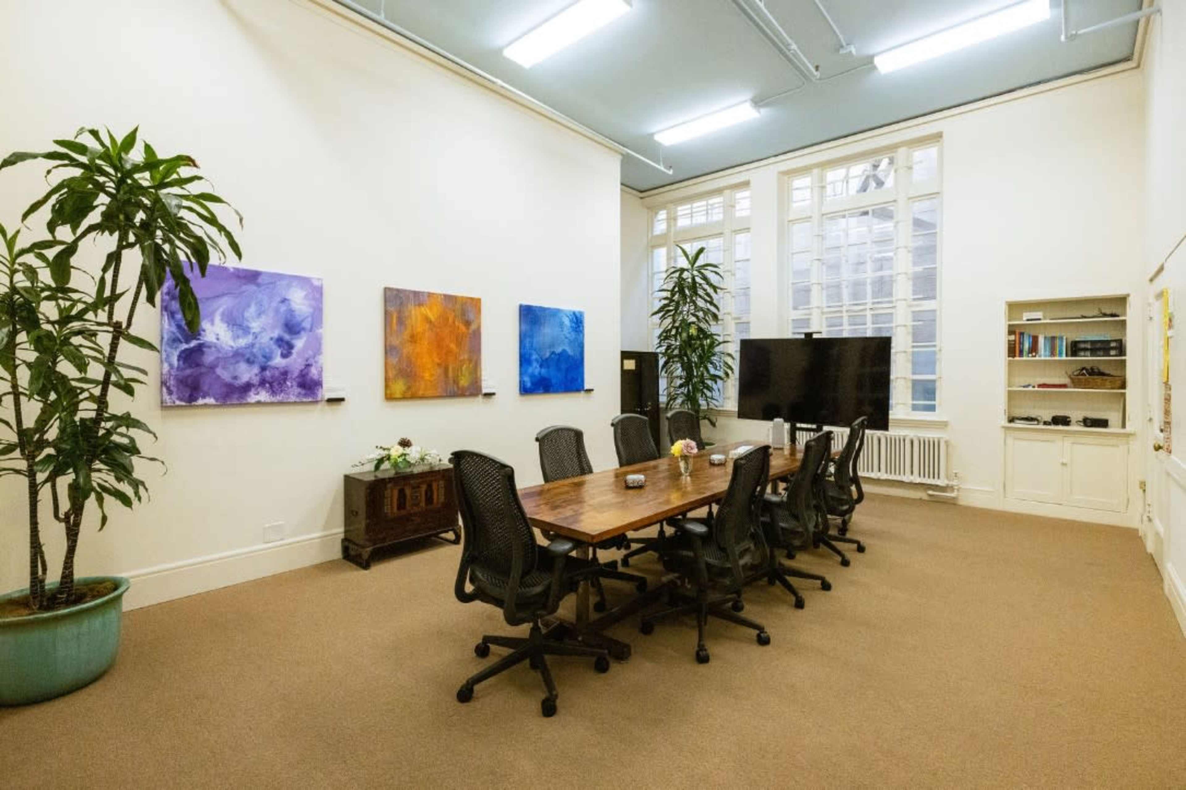 A conference room features a long wooden table surrounded by black office chairs, with large windows and colorful abstract paintings on the walls.