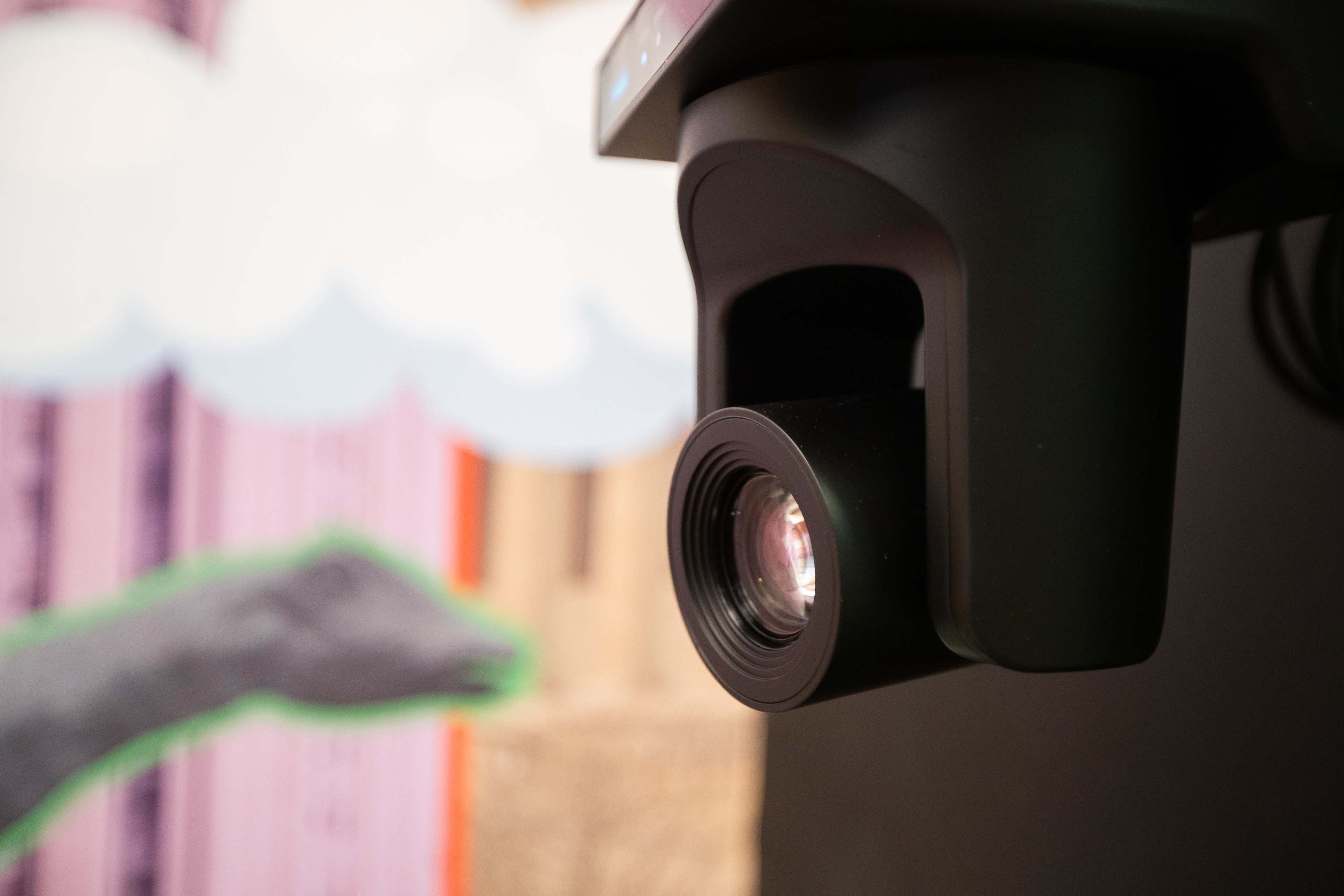 A close-up of a black camera lens mounted on a device, with a colorful background featuring stylized clouds and a hand.