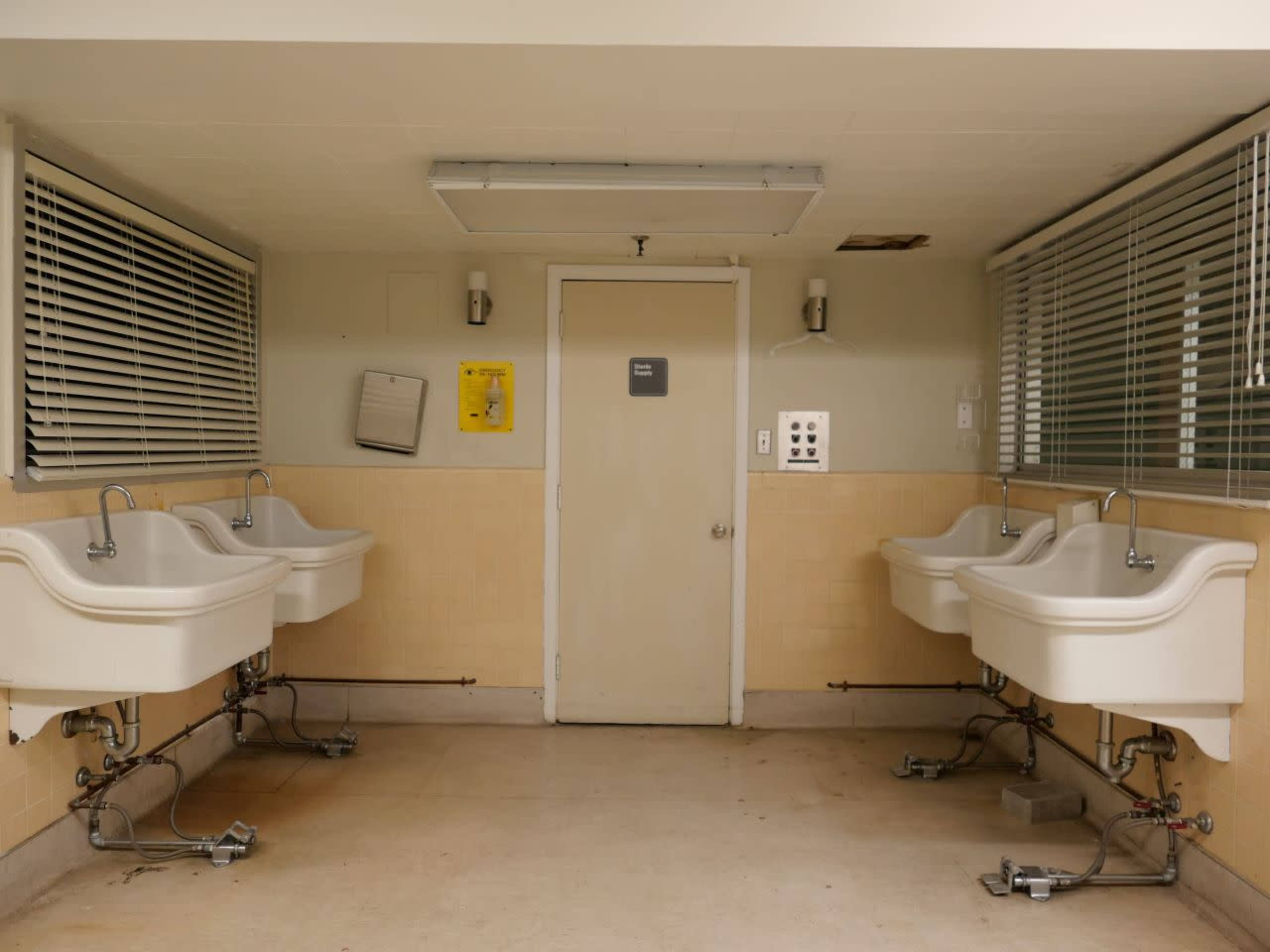 A room with four utility sinks arranged along two walls, facing a closed door marked with a sign.