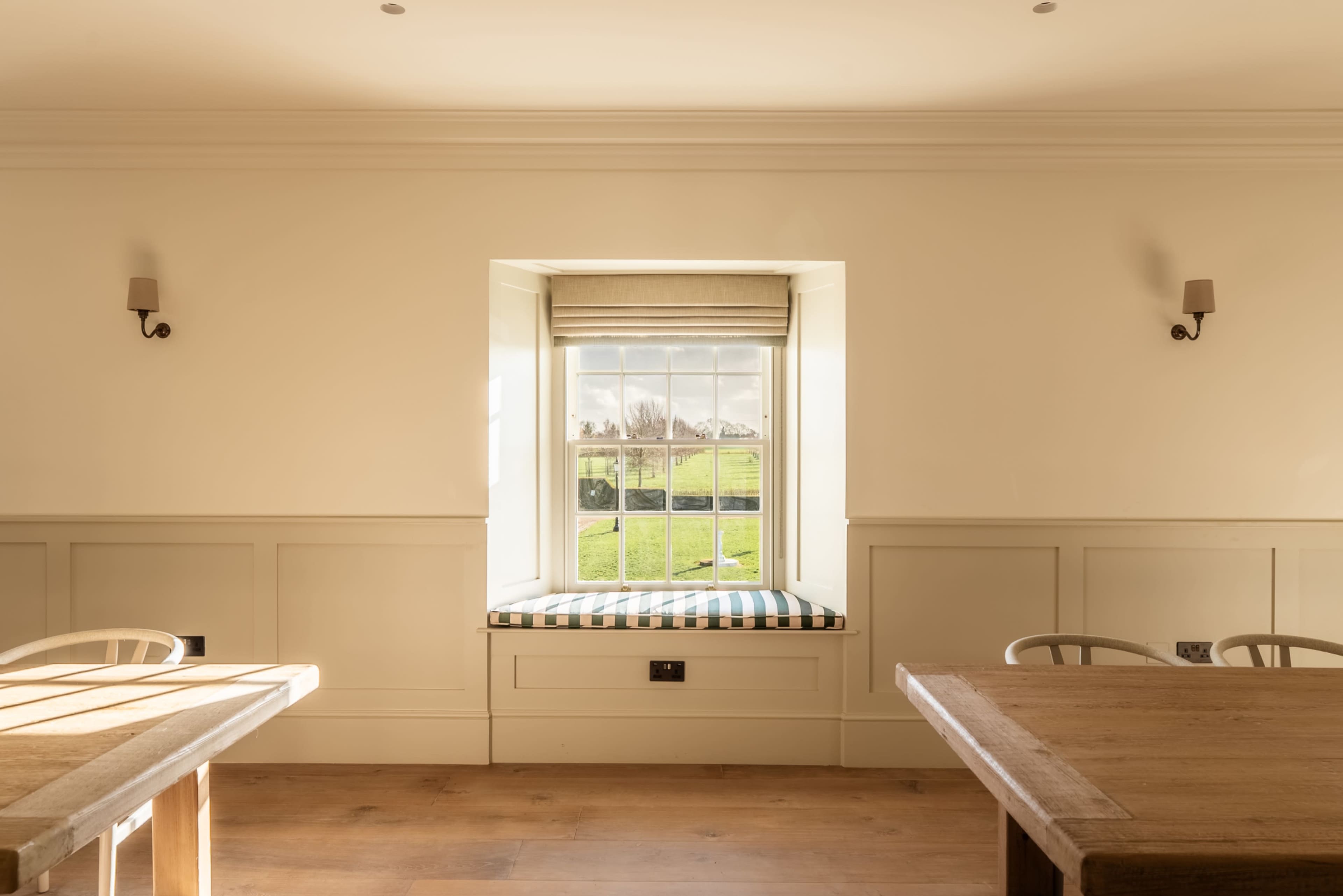 A bright room features a window seat beneath a large window with a view of the outdoors, surrounded by wooden tables and light-colored walls.