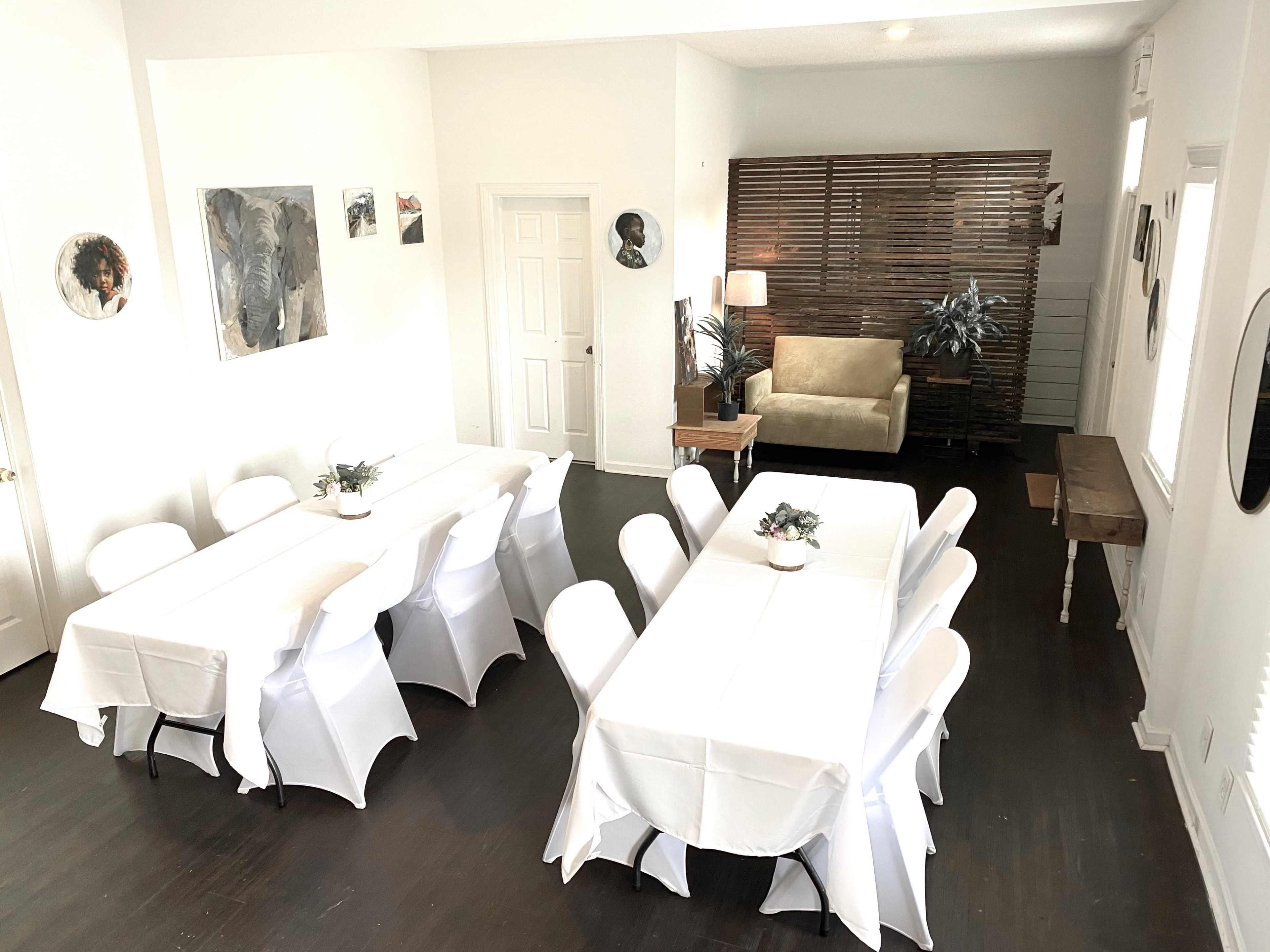A spacious room with two neatly arranged tables covered with white tablecloths, surrounded by chairs, and a cozy seating area against a wooden accent wall.