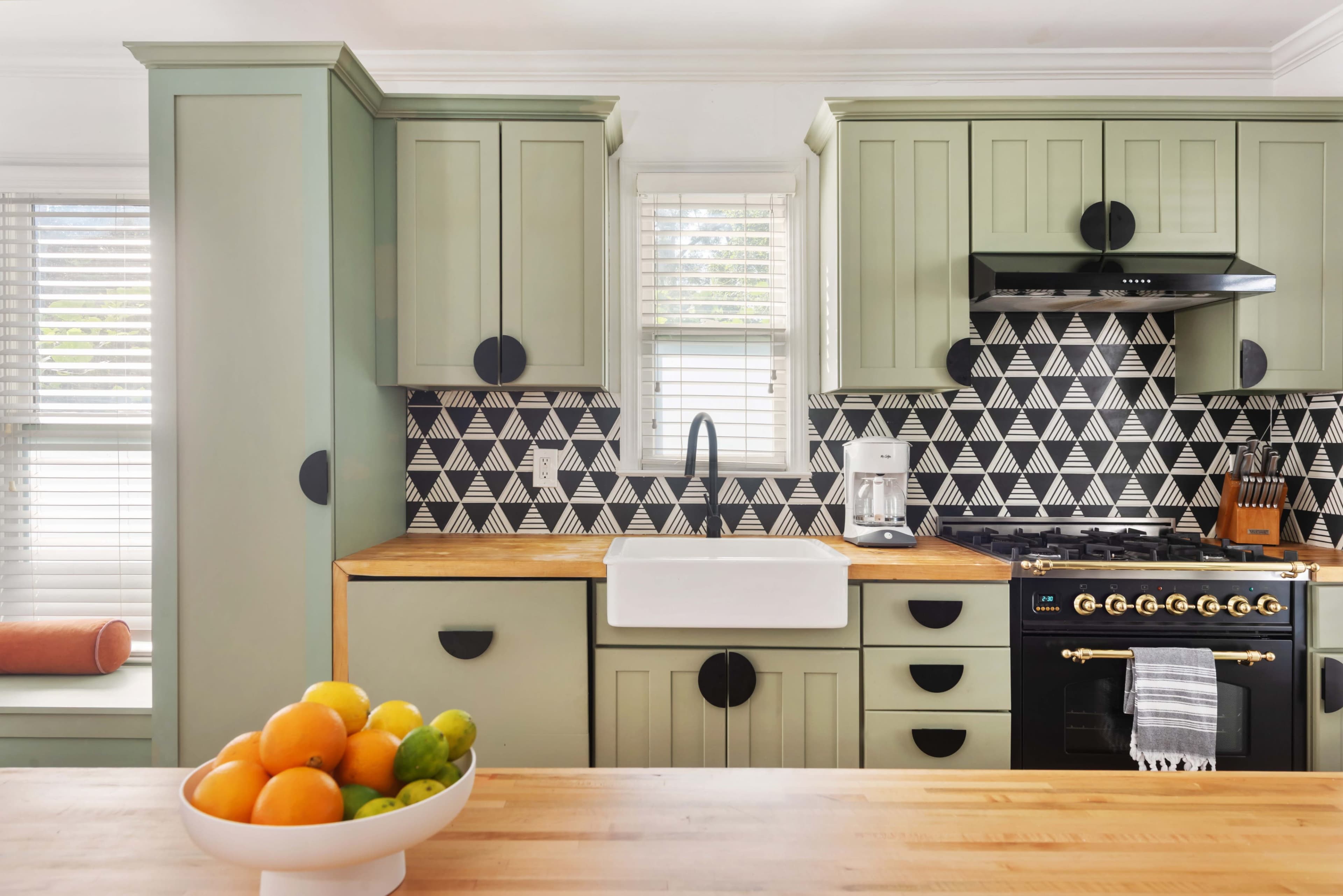 The kitchen features green cabinetry, a farmhouse sink, and a black and white geometric backsplash, with a wooden countertop and a bowl of colorful fruit on the island.