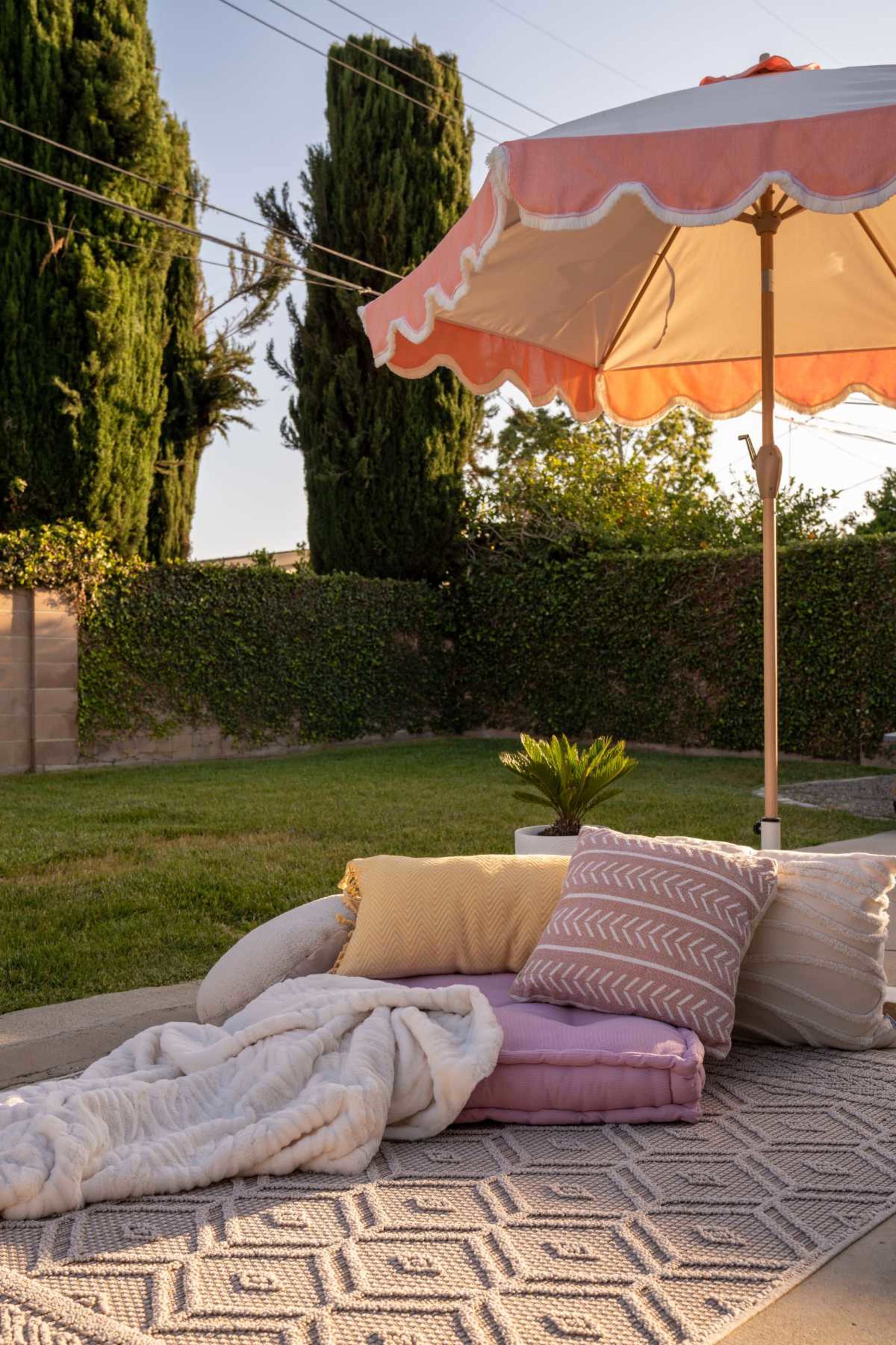 A patio scene features a large parasol over a patterned rug, with layered cushions and a soft throw blanket, set against a backdrop of tall greenery.