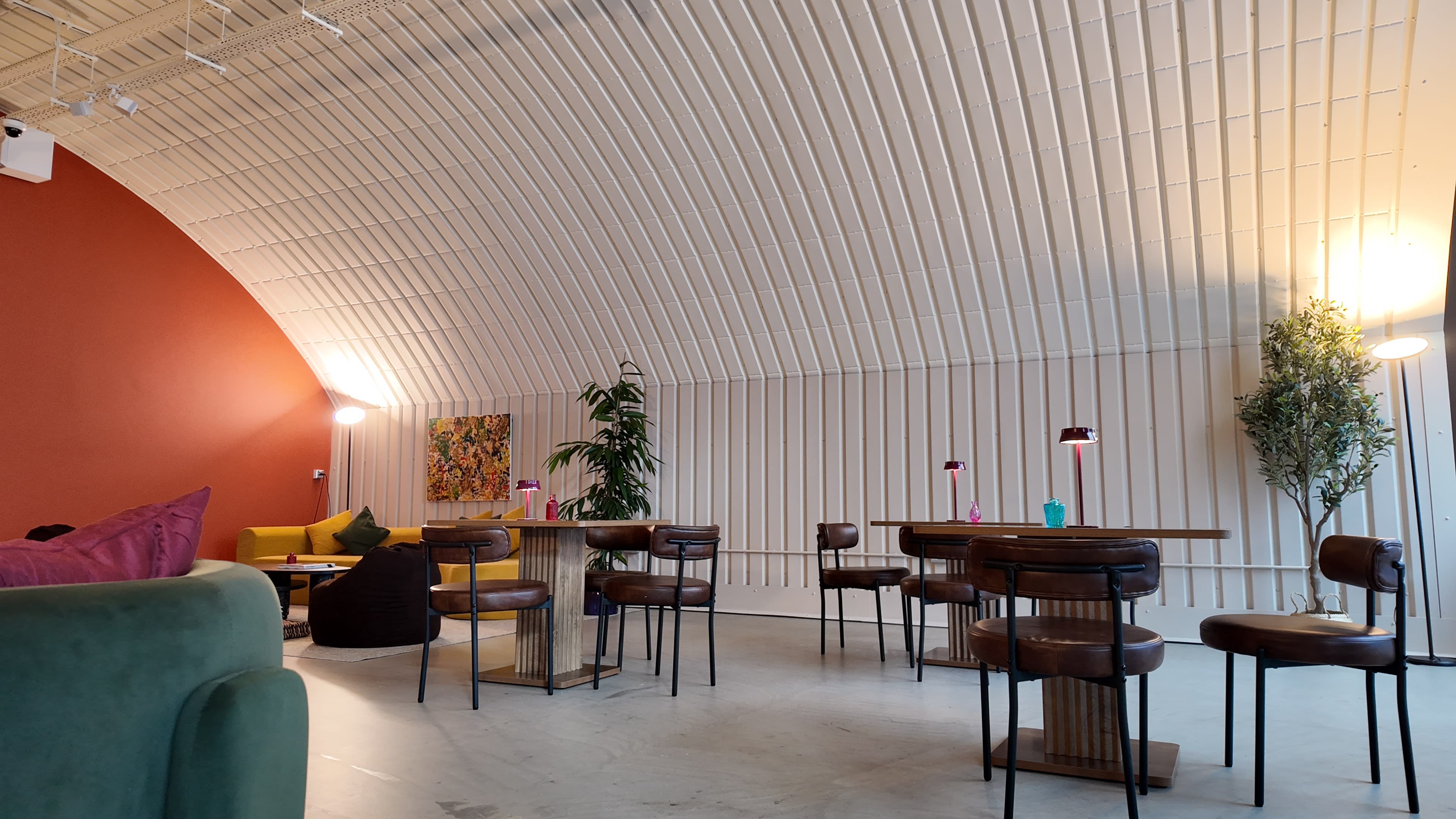 The image shows a modern cafe interior with curved white walls, wooden tables, and a mix of seating arrangements, including chairs and a green sofa.