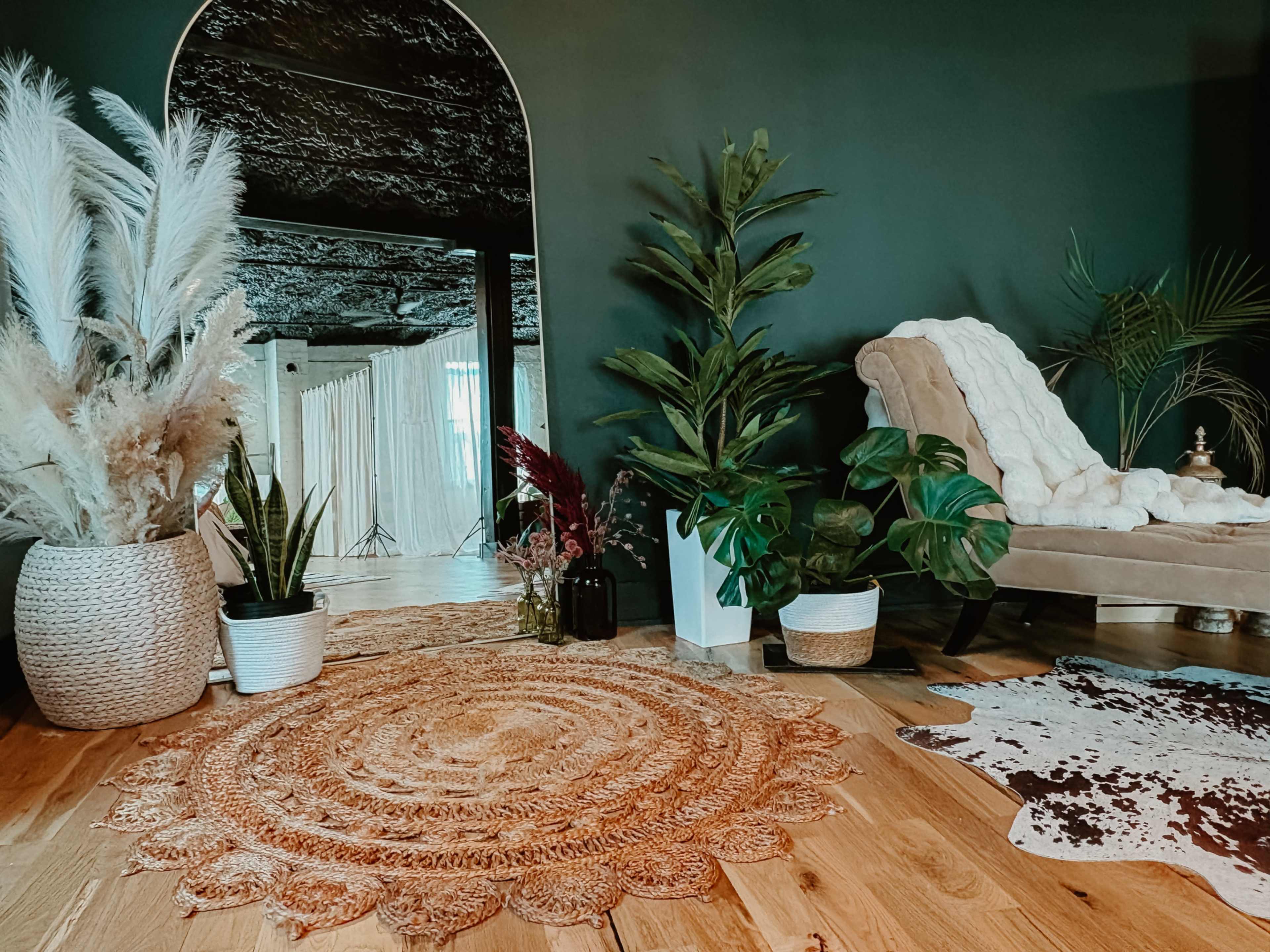 The image shows a cozy interior space featuring a textured rug, various plants in decorative pots, a comfortable chair, and a large mirror reflecting soft lighting.