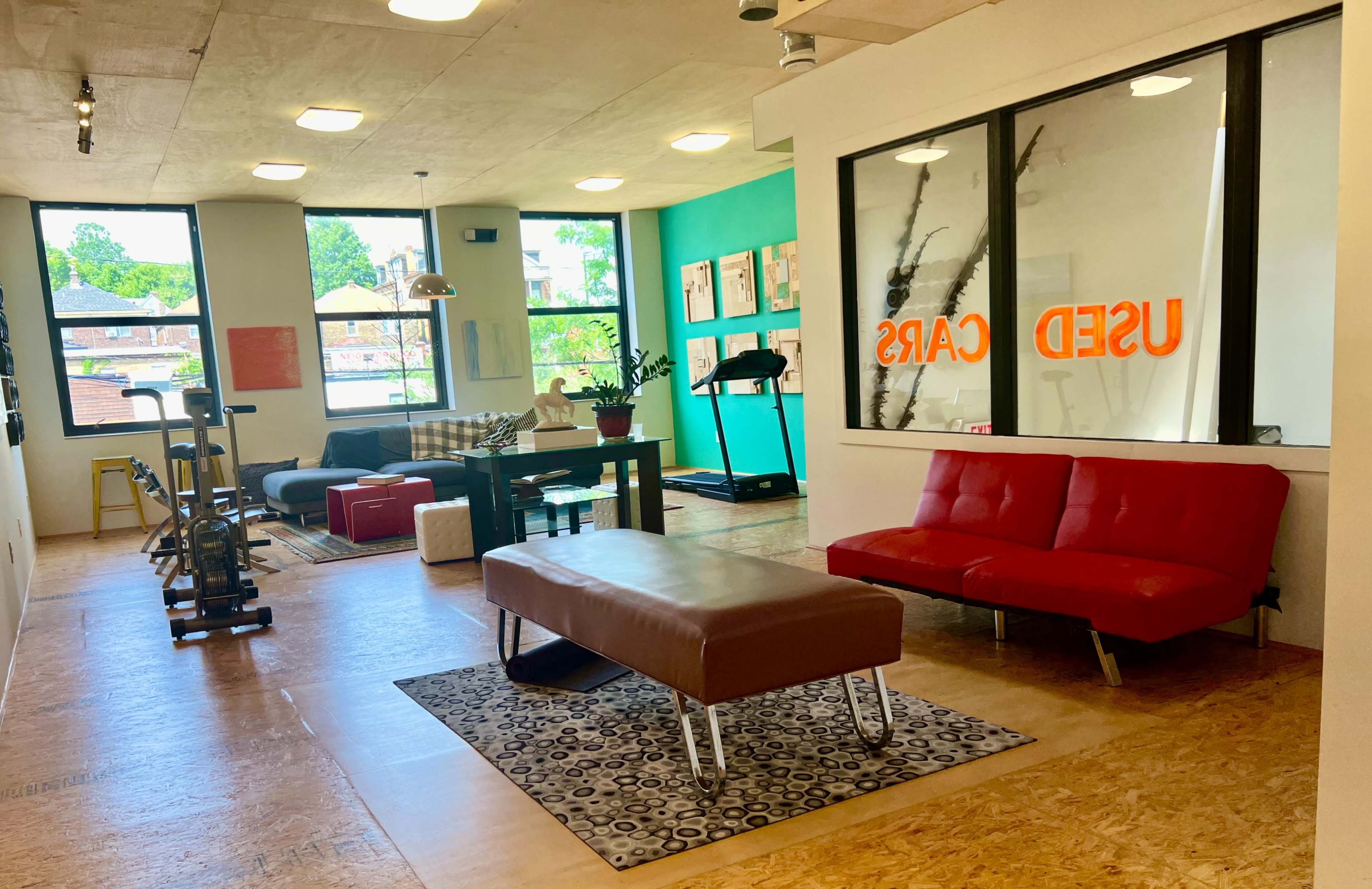 A spacious, modern workspace with a red couch, a wooden table, exercise equipment, and large windows displaying the words "USED CARS" in bright orange.