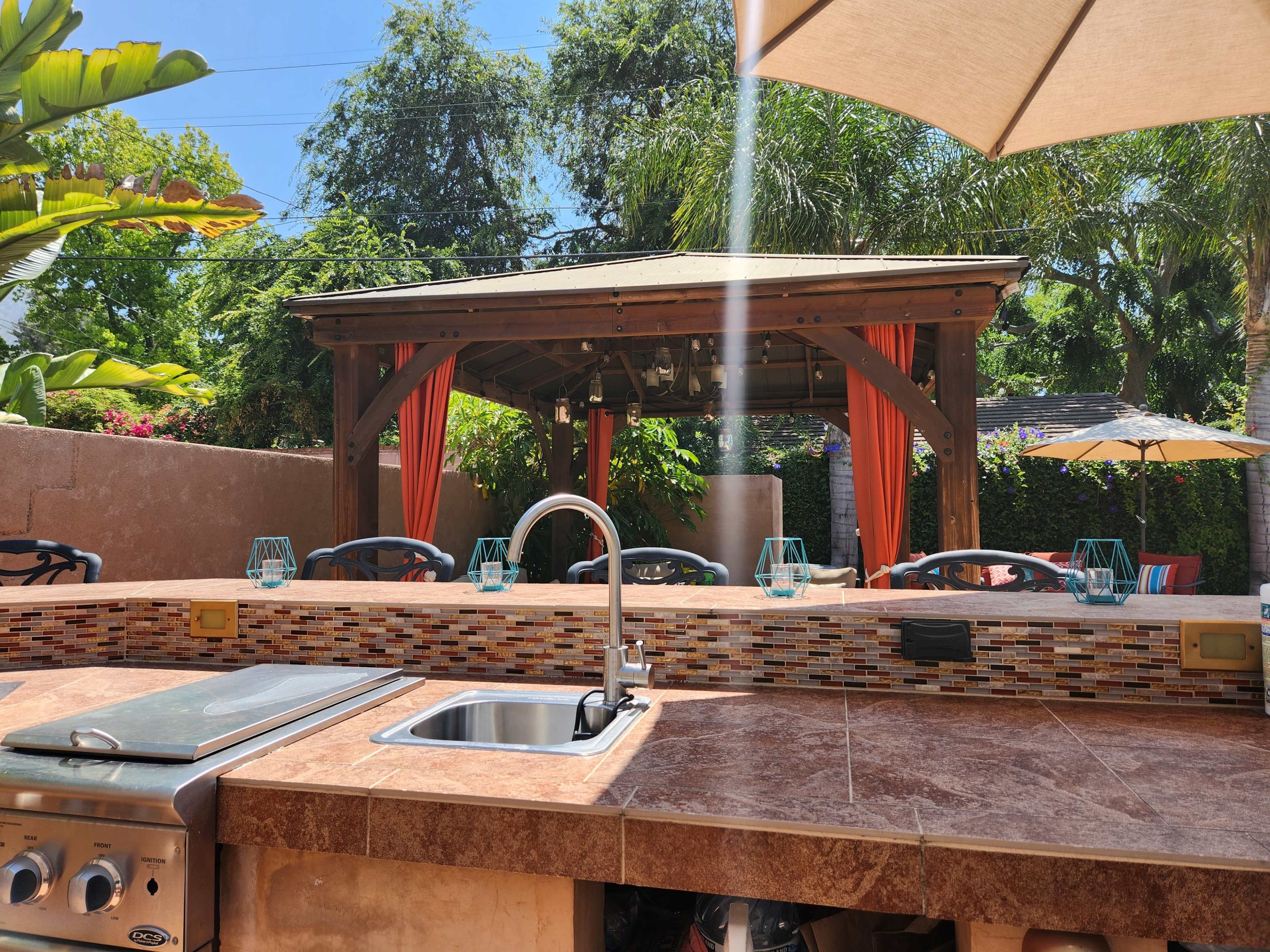 The image shows an outdoor kitchen area overlooking a covered seating space surrounded by greenery and colorful decor.