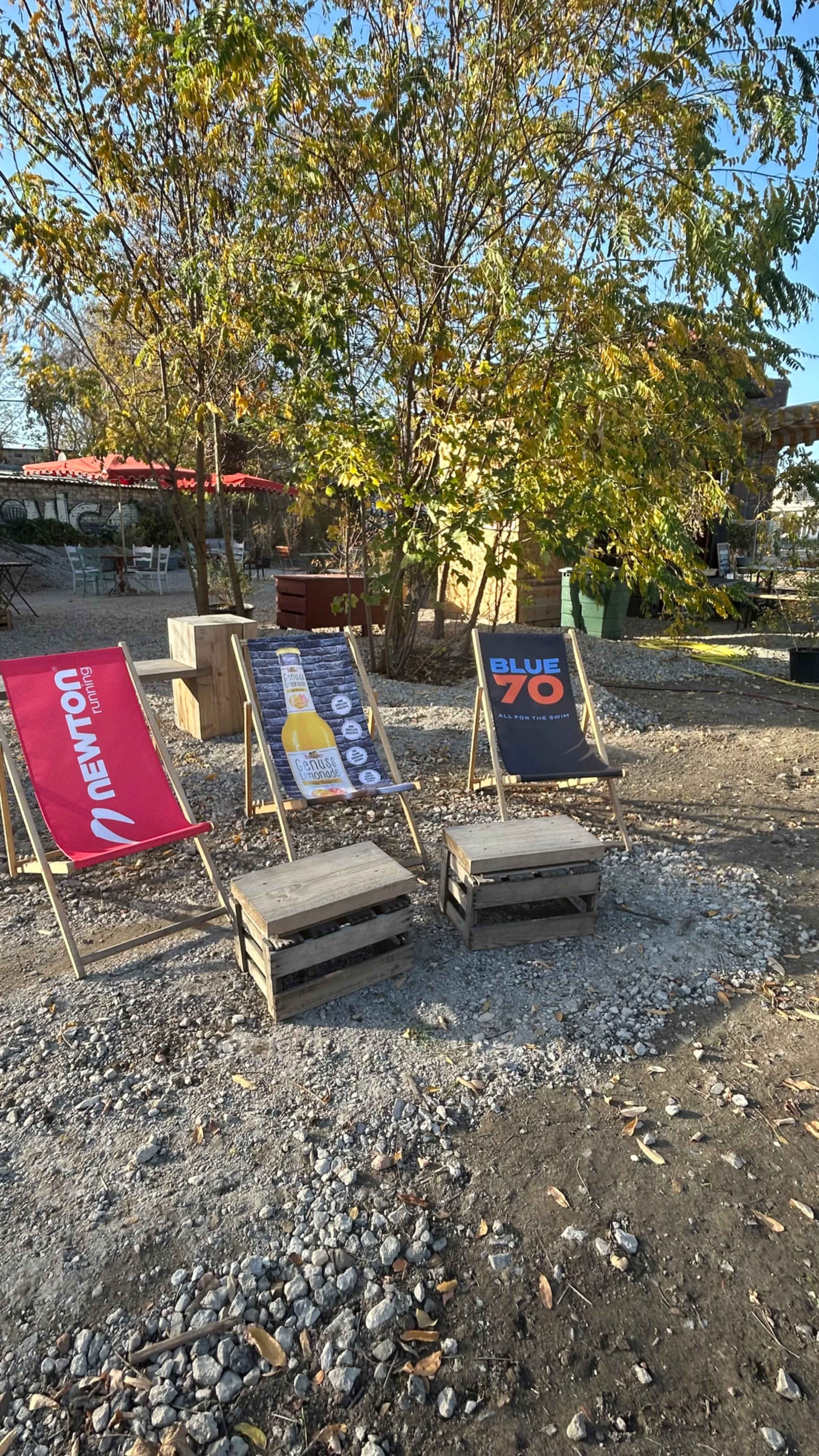 The image shows three wooden lounge chairs with different branded advertisements, positioned on gravel under trees in a recreational outdoor setting.