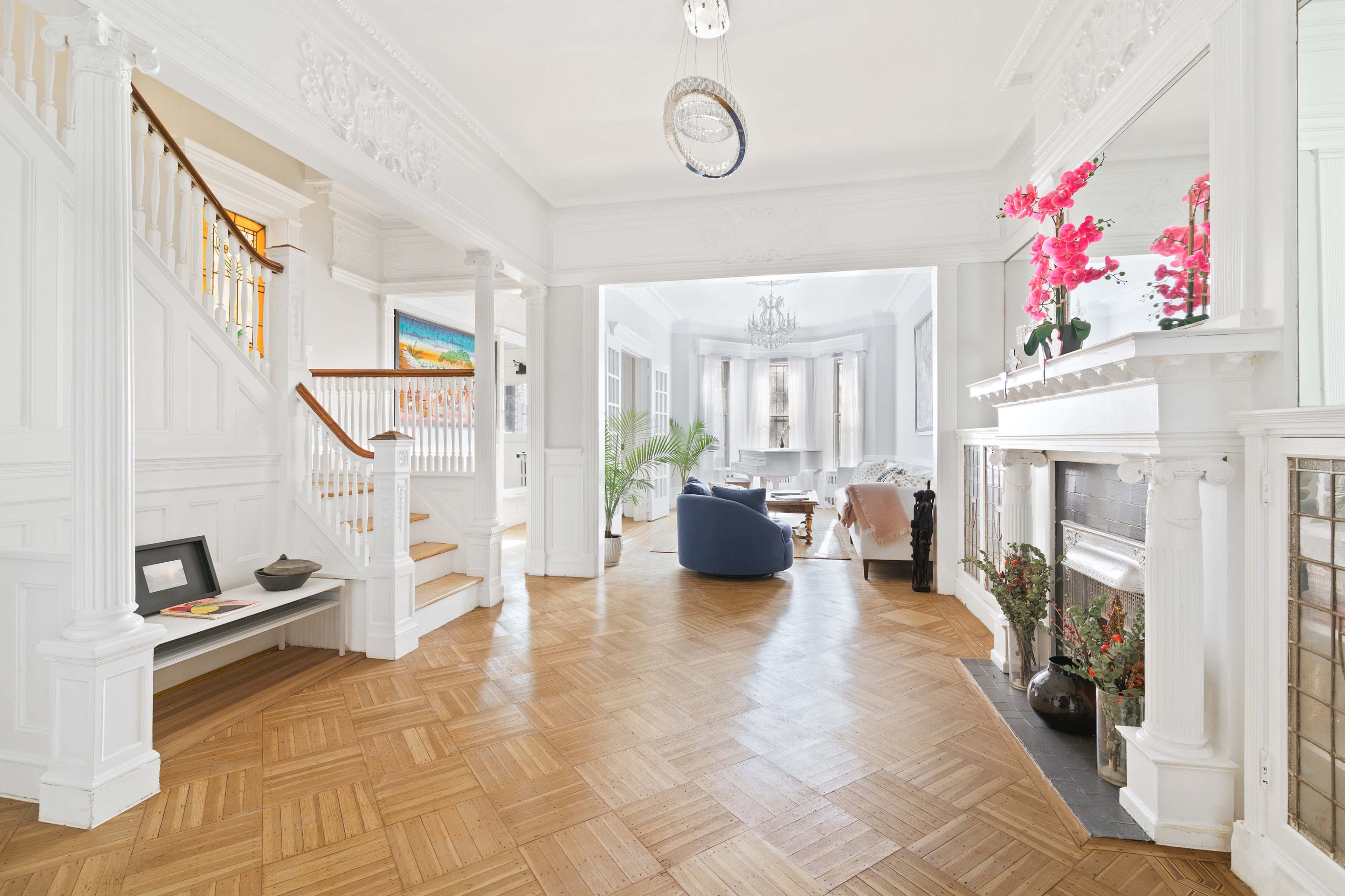 A bright entryway with a wooden floor, an elegant staircase, and a view into a spacious living room featuring a modern seating area and decorative plants.