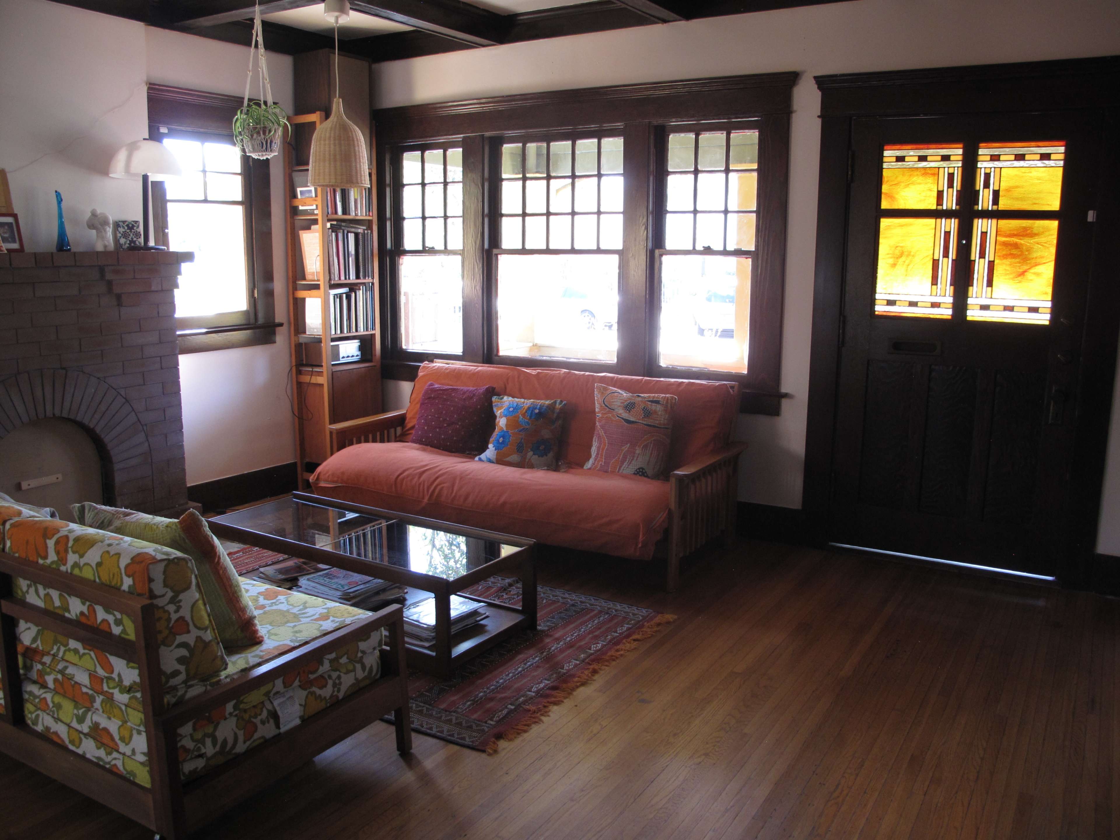The image shows a living room featuring a sofa and armchair, a coffee table, a fireplace, and large windows with stained glass.