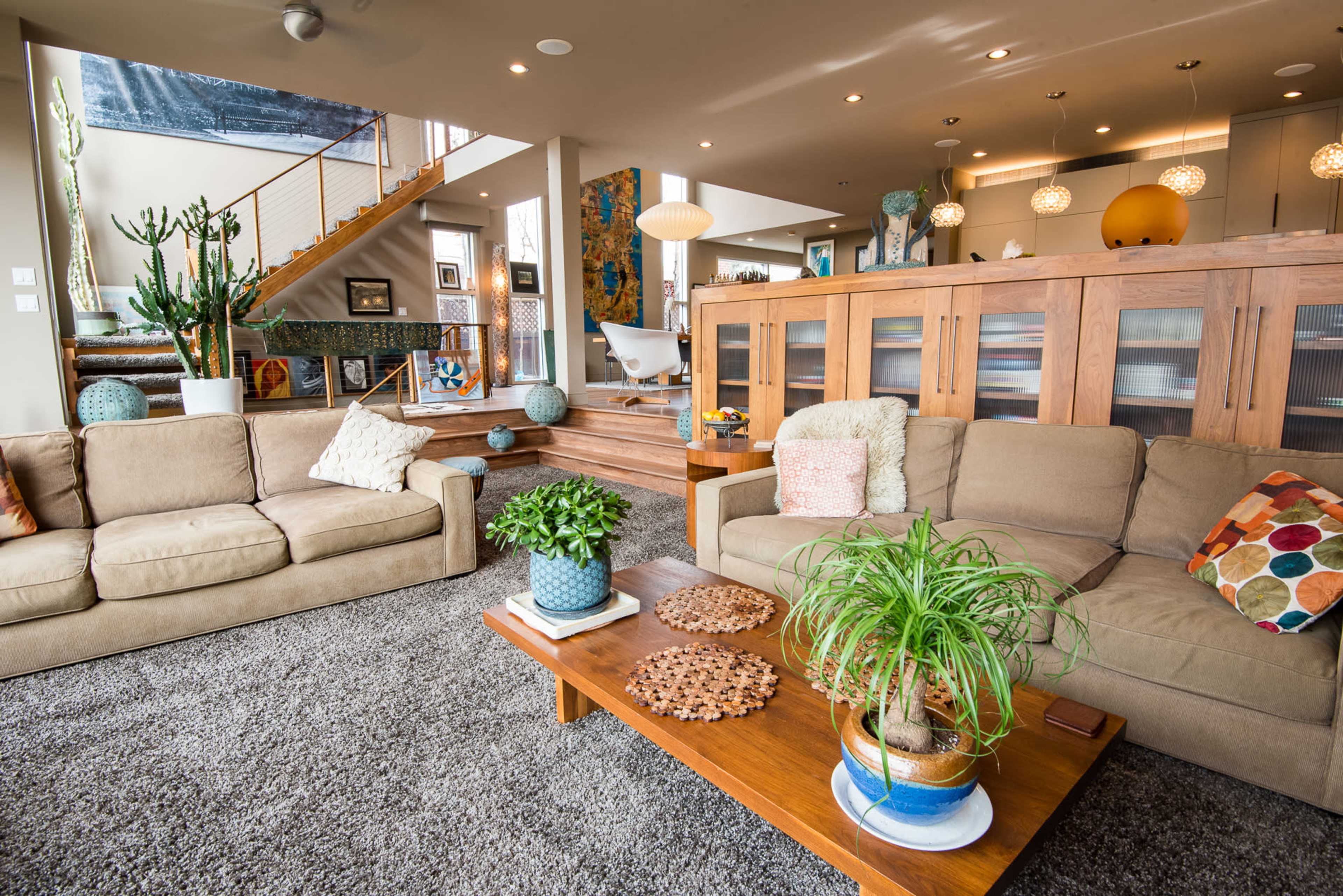 The image depicts a modern living room with beige sofas, a wooden coffee table, and a staircase leading to an upper level, surrounded by various plants and colorful decor.