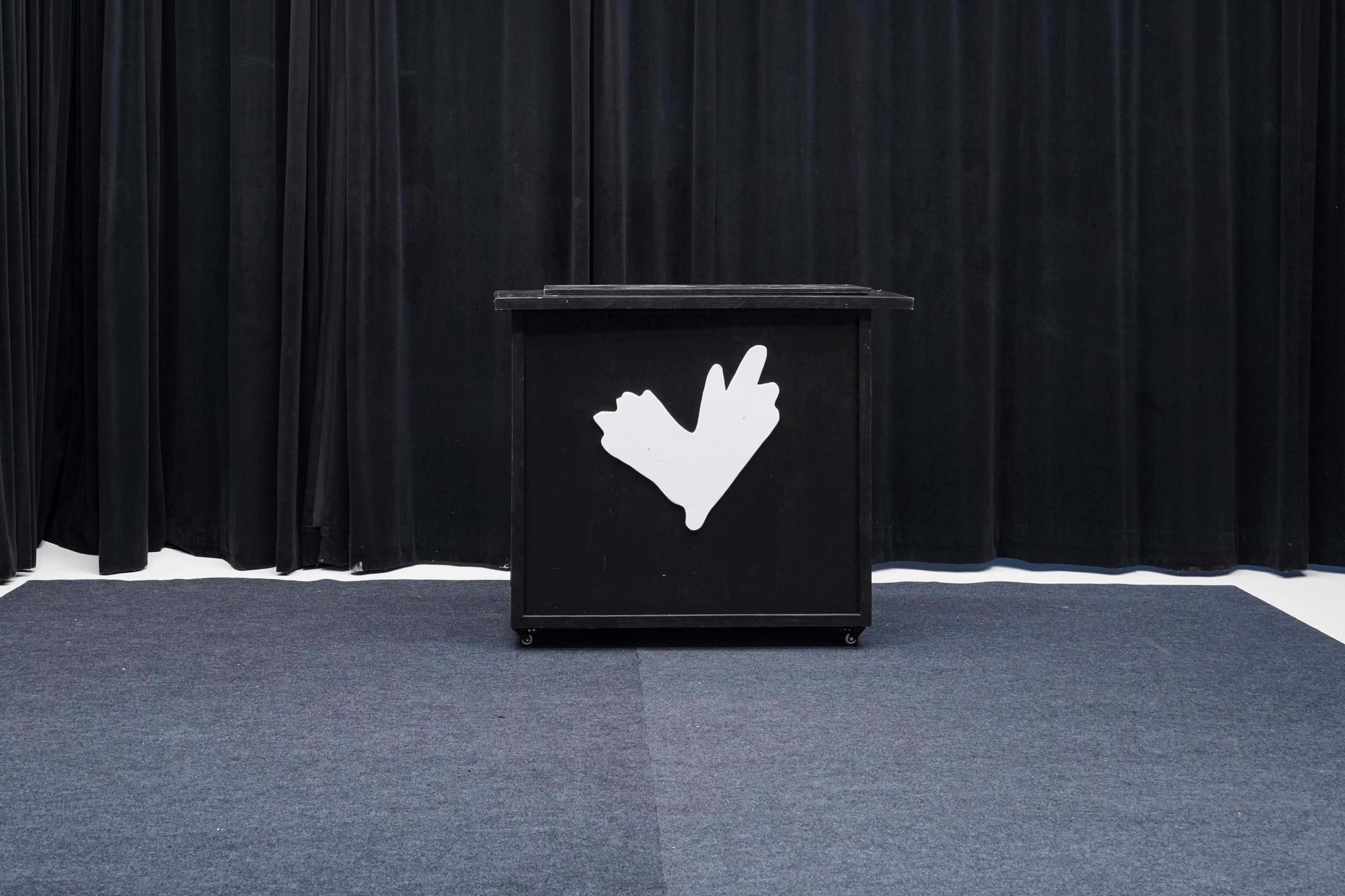 A black podium with a white hand symbol is positioned in front of a black curtain on a blue carpeted floor.
