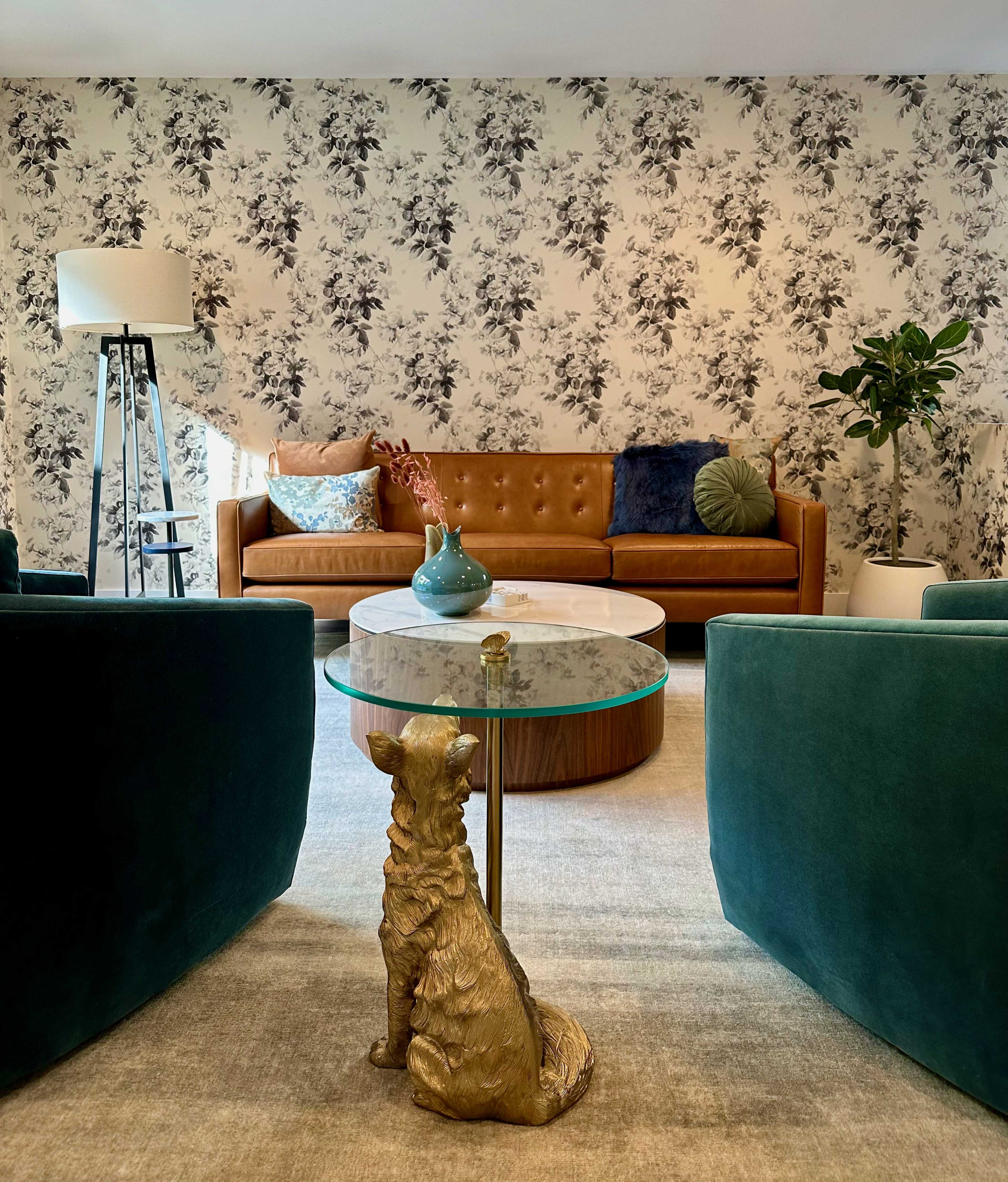 The image shows a living room with a brown leather sofa, two teal armchairs, a round coffee table with a floral base, and a decorative dog sculpture beside the table, all set against a floral wallpaper backdrop.