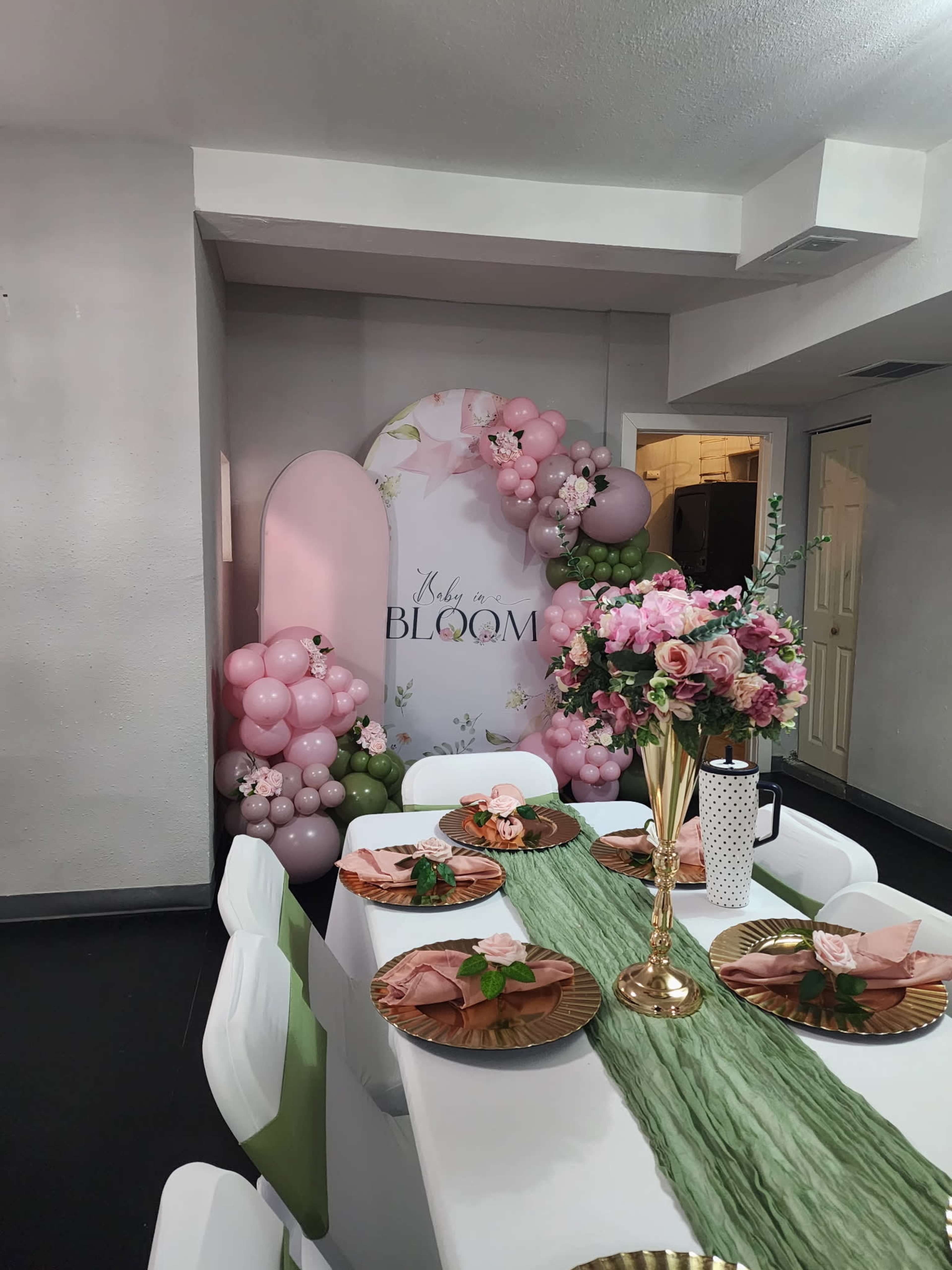 A dining table is set with pink plates and flowers, beside a decorative backdrop featuring balloons and a "Baby in Bloom" theme.