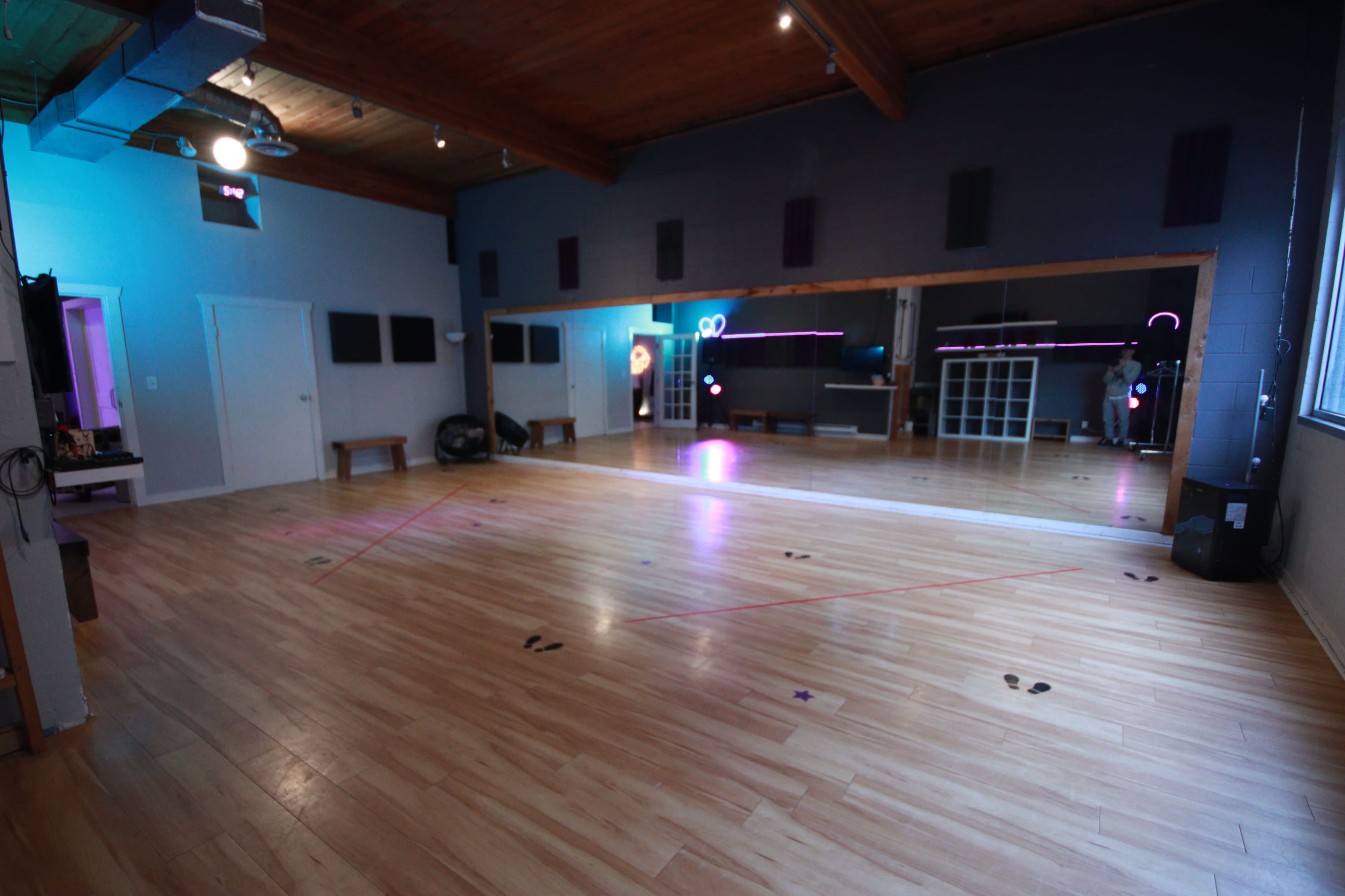 A spacious dance studio with wooden flooring, wall-mounted speakers, and a large mirror reflecting colorful lights.