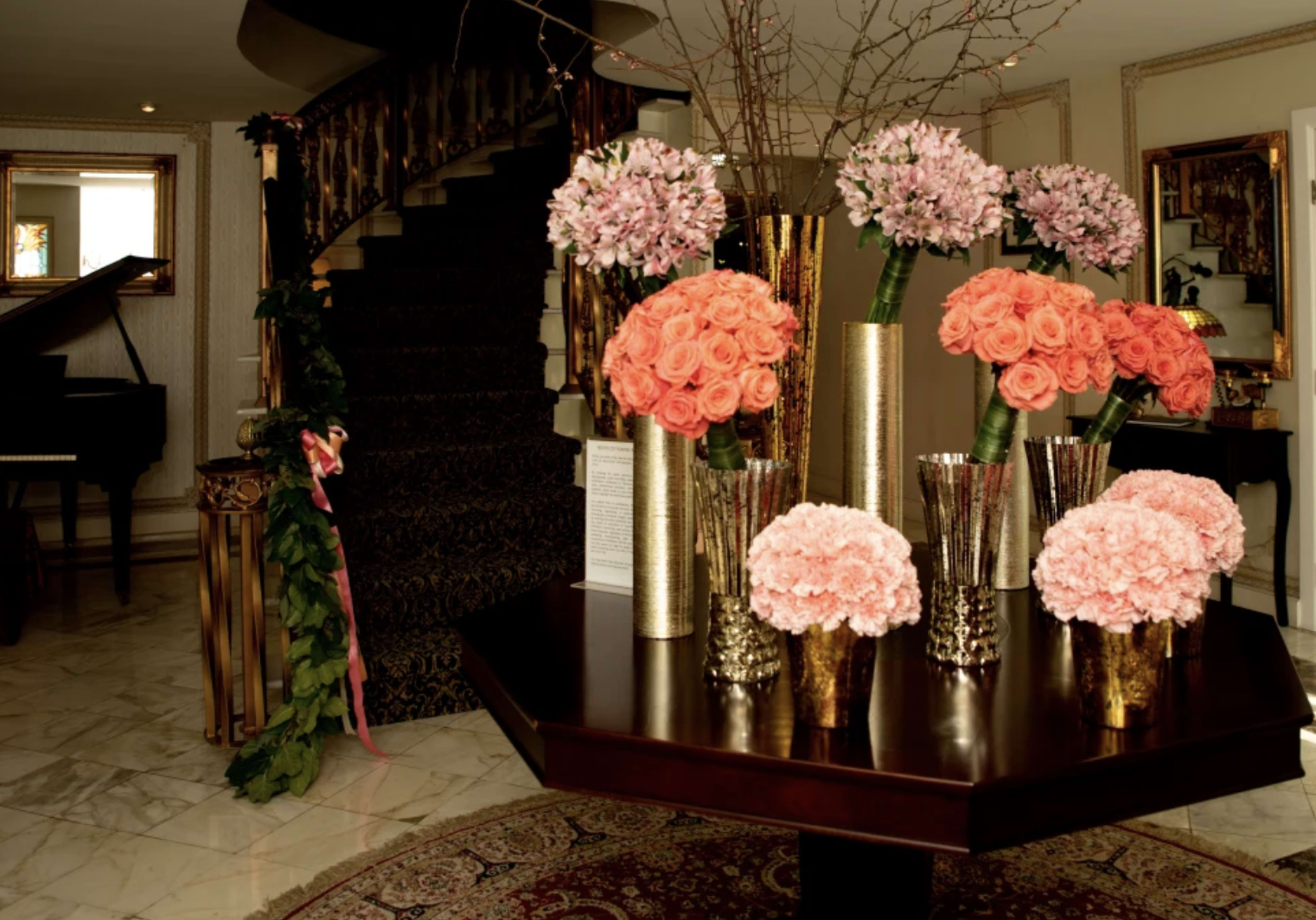 A foyer features a round wooden table with vibrant flower arrangements in metallic vases and a staircase in the background.