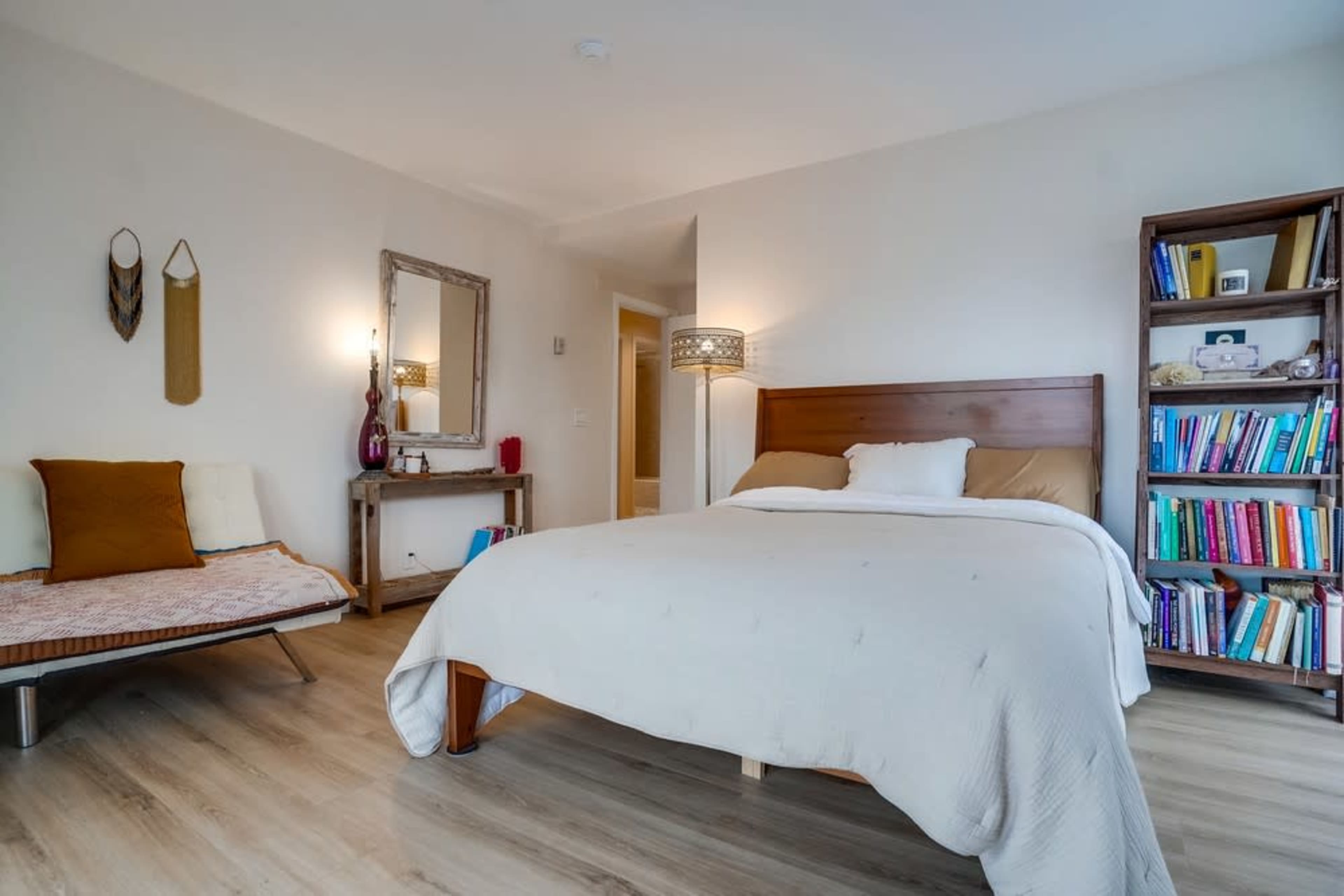 The bedroom features a bed with a light-colored duvet, a bookshelf filled with books, and a seating area with a small chair and a table.