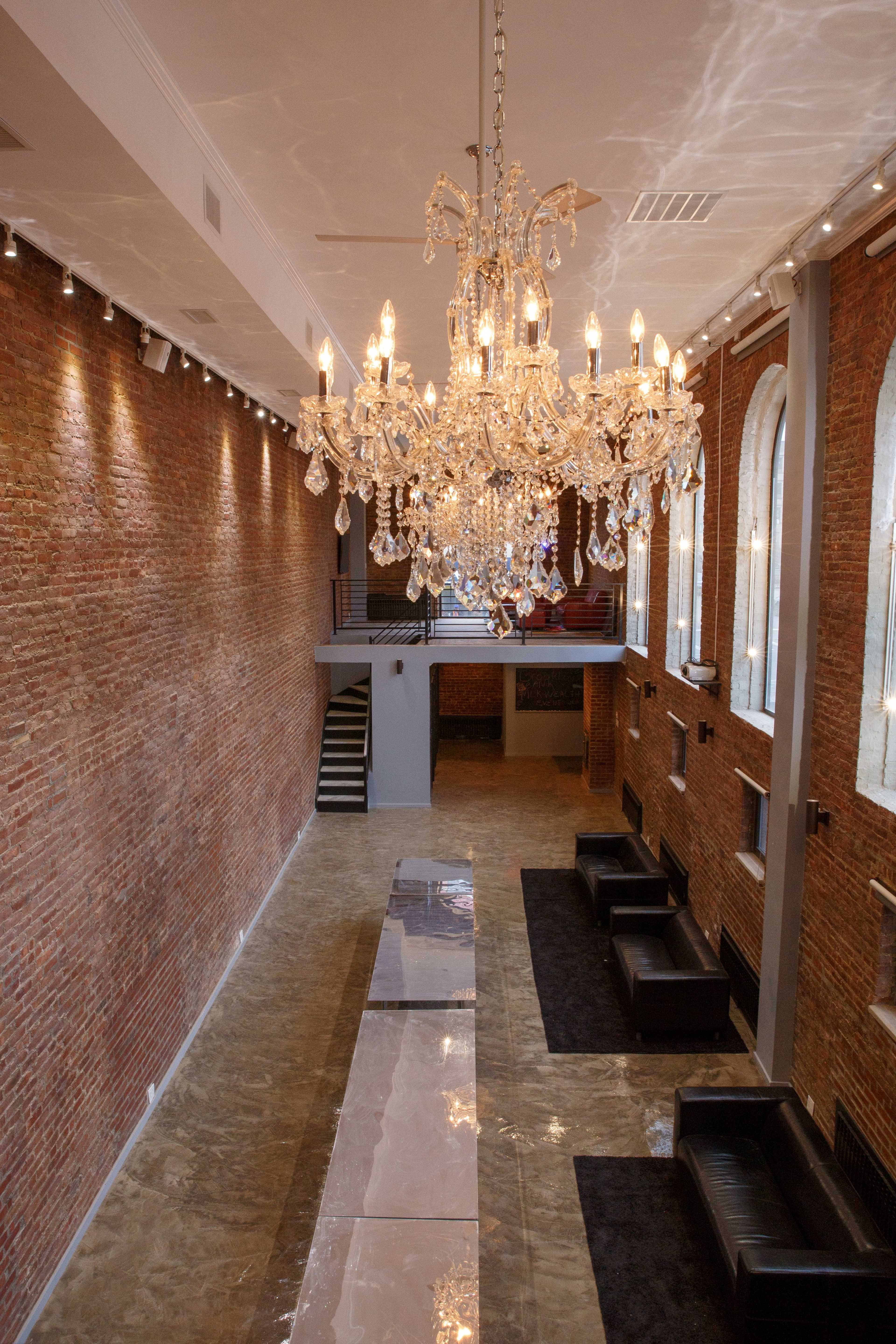 A spacious interior features a large chandelier suspended from the ceiling, with exposed brick walls and a polished floor, complemented by black sofas along the sides.
