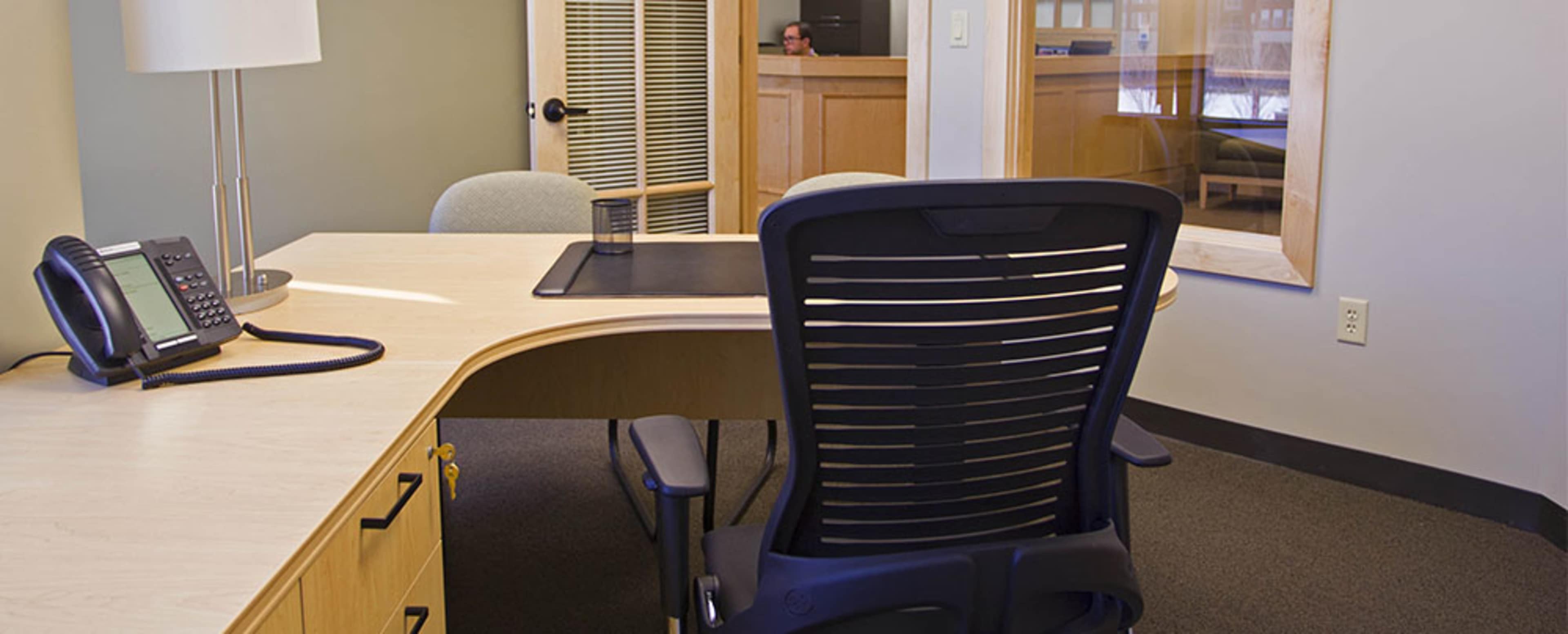 The image shows an empty office with a curved desk, a black ergonomic chair, a telephone, and a background area with a person visible through a glass partition.