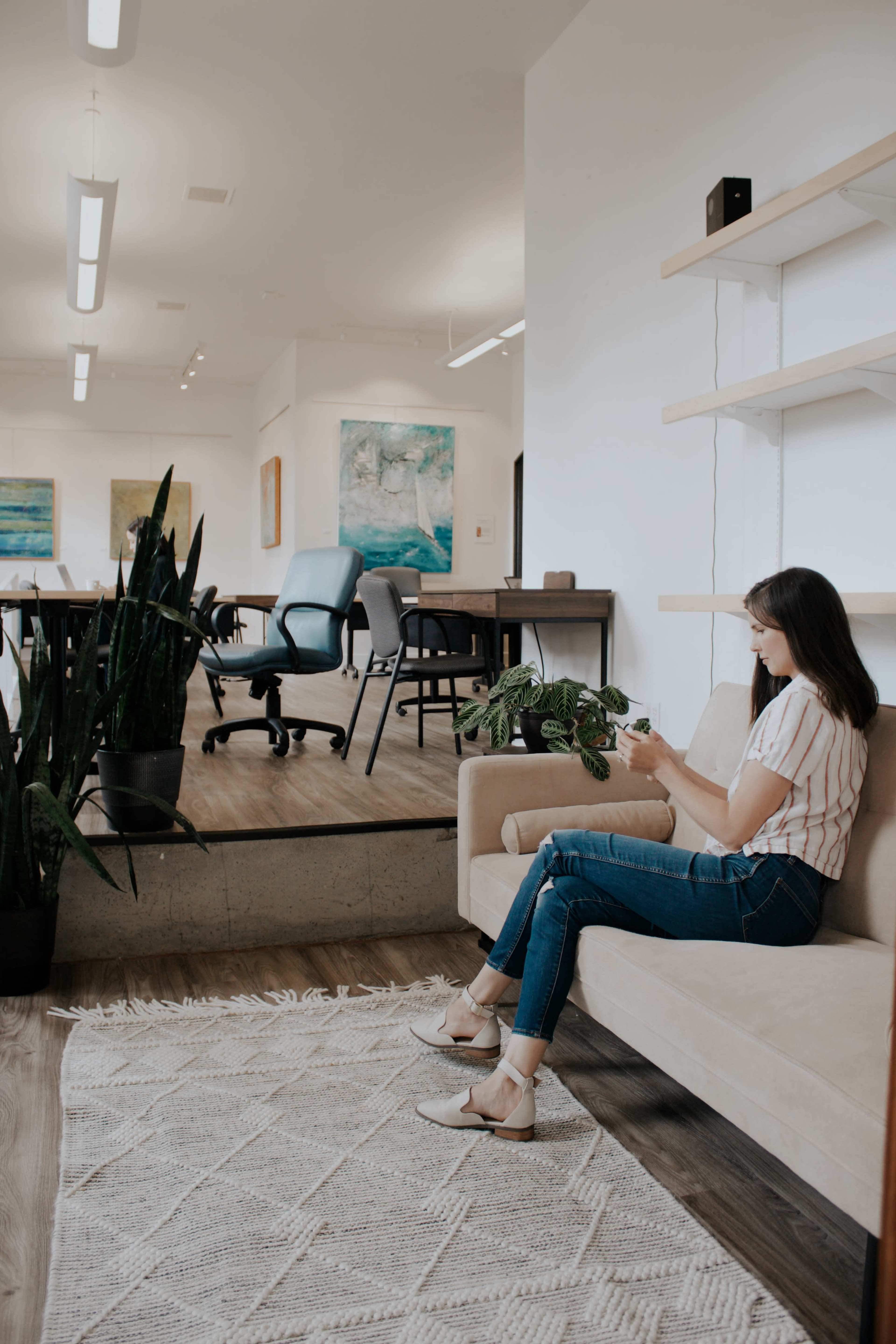A woman sits on a beige sofa in a bright, modern office space, engaging with her phone while surrounded by plants and contemporary furniture.