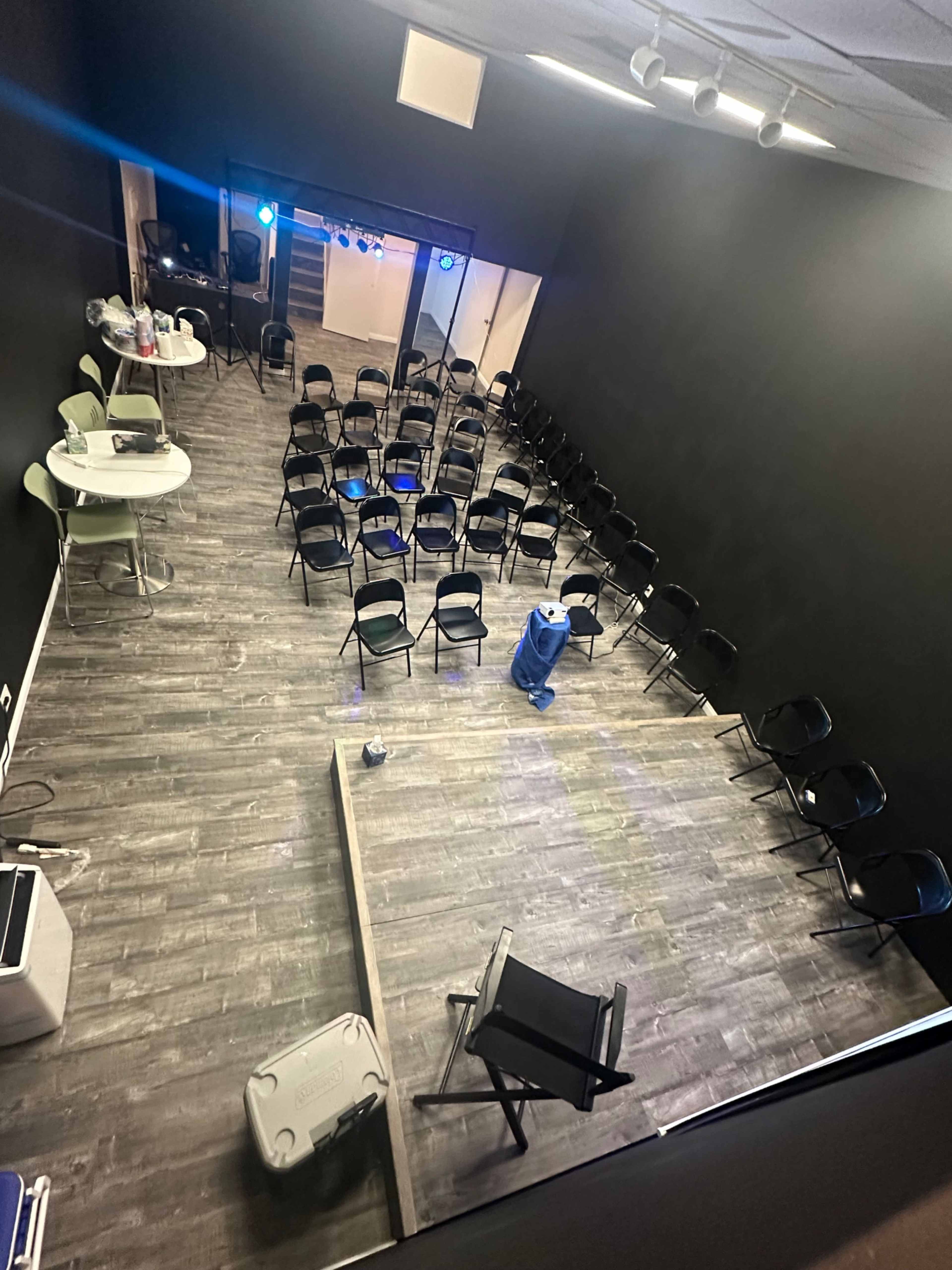 The image shows a spacious room with a black wall, featuring rows of chairs arranged for an event, with a small stage area and equipment at the front.