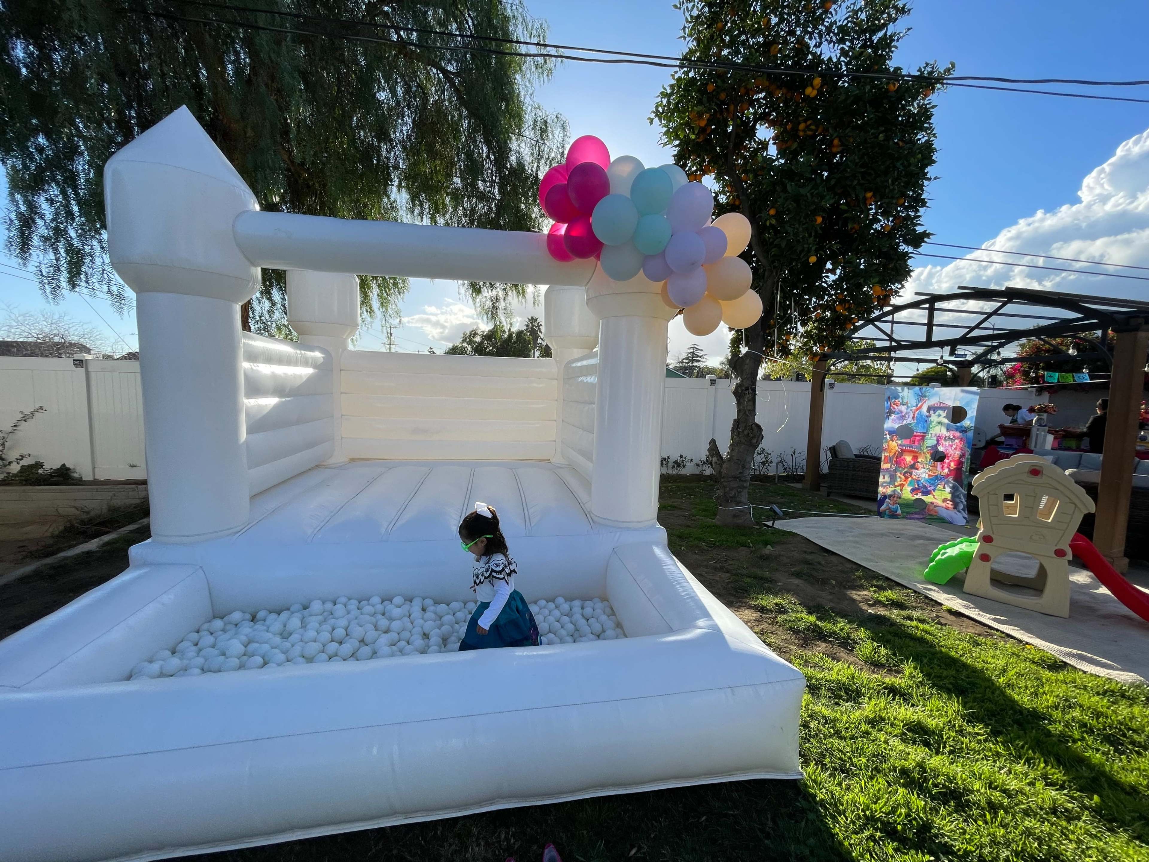 A child stands inside a white inflatable bounce house filled with colorful balls, while balloons cluster at one corner and a playground structure is visible in the background.