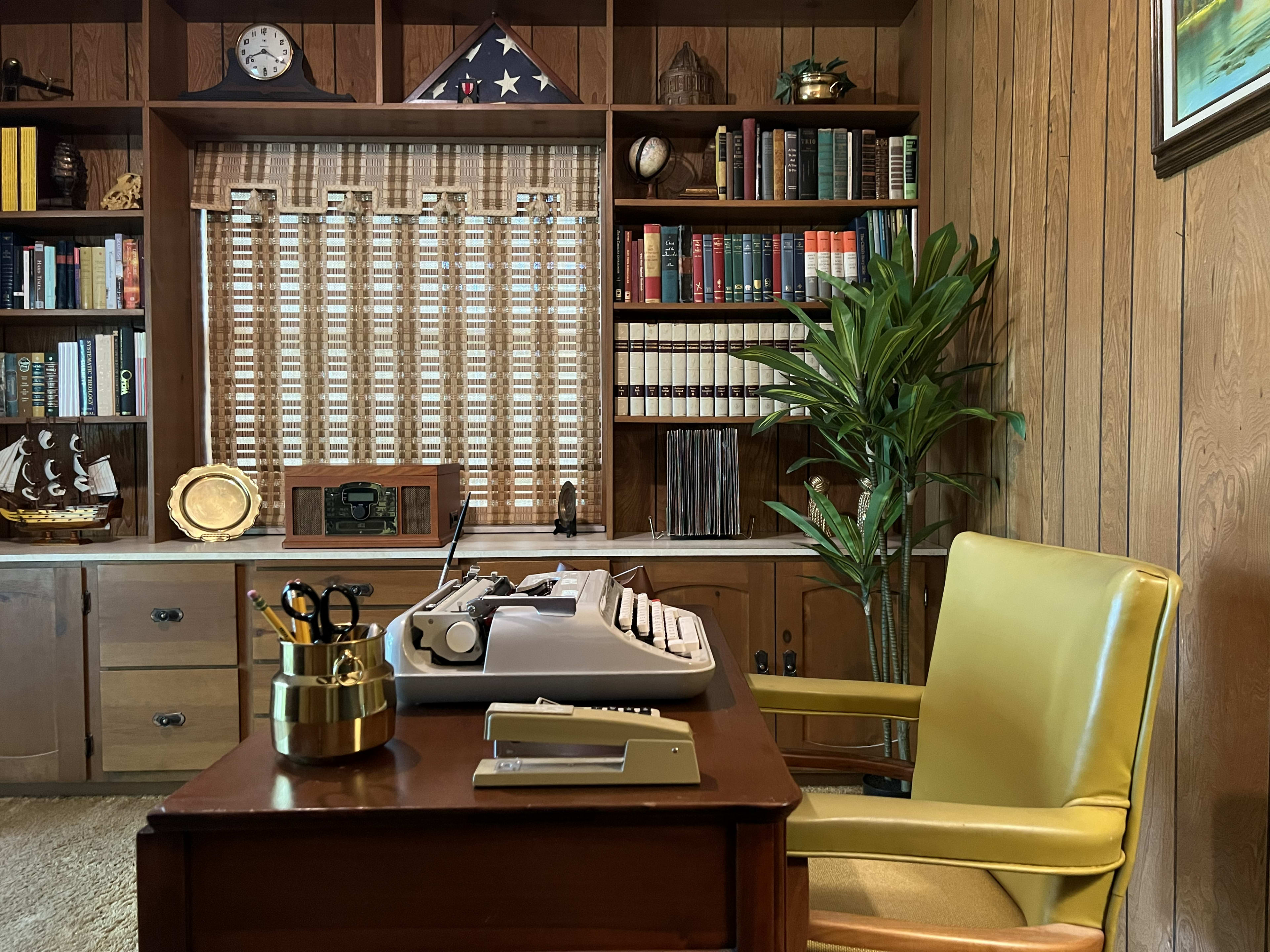 The image shows a wooden office with a desk, a typewriter, a yellow chair, bookshelves filled with books, and a window with closed blinds.