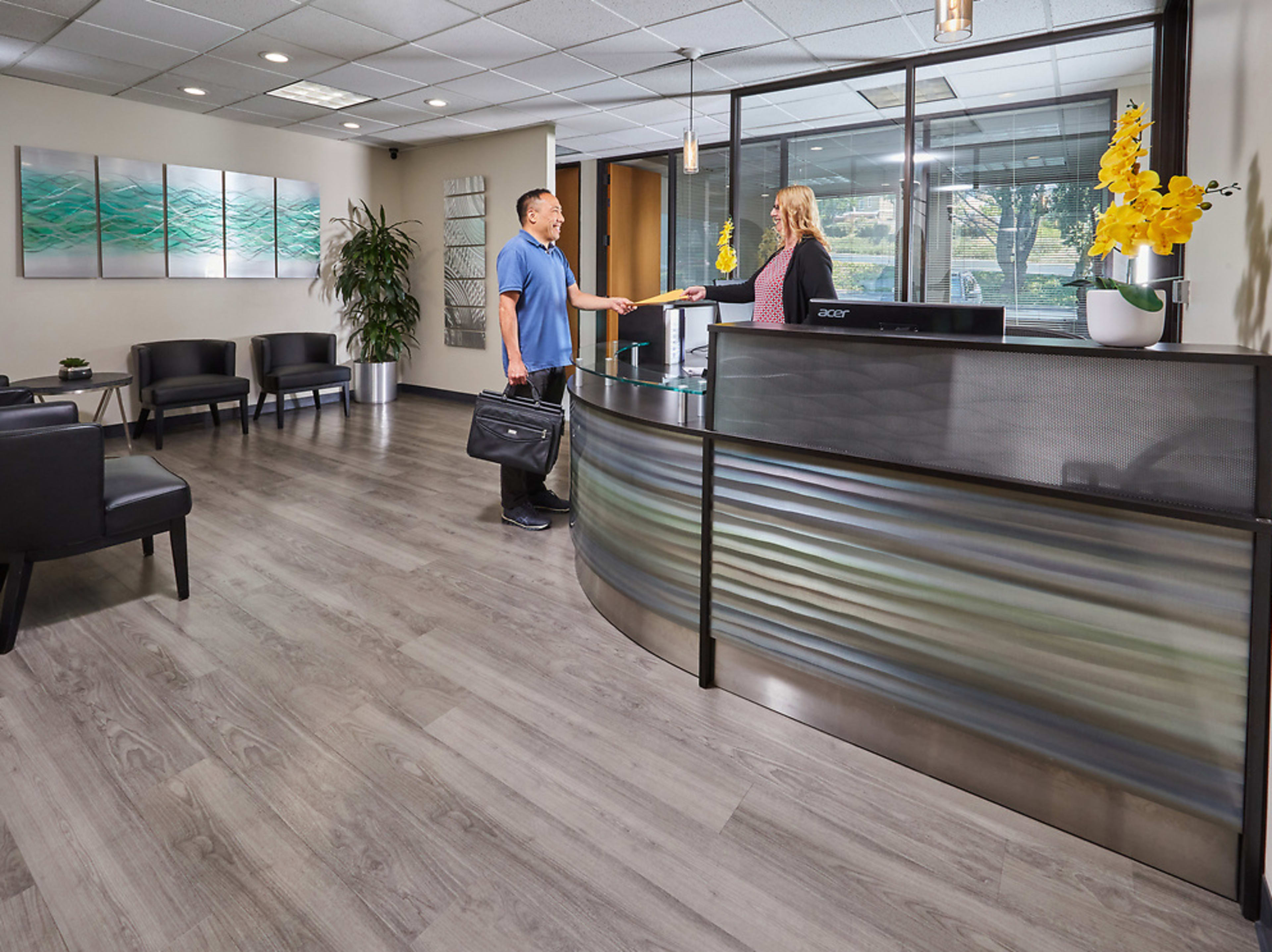A man in a blue shirt hands a document to a receptionist at a modern office reception area with gray flooring and decorative plants.